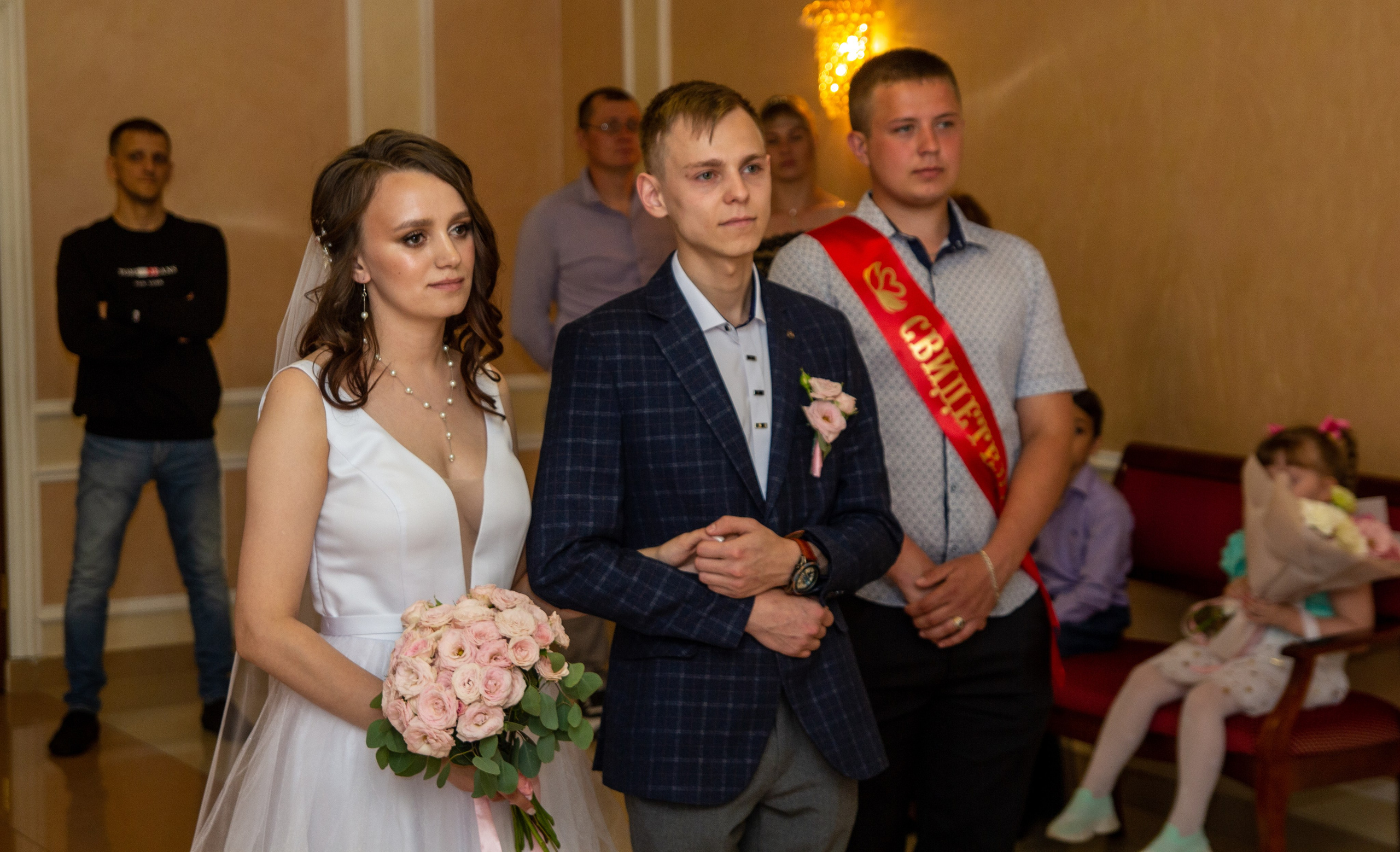 Жених и невеста стоят с гостями на торжественной церемонии — невеста держит букет.