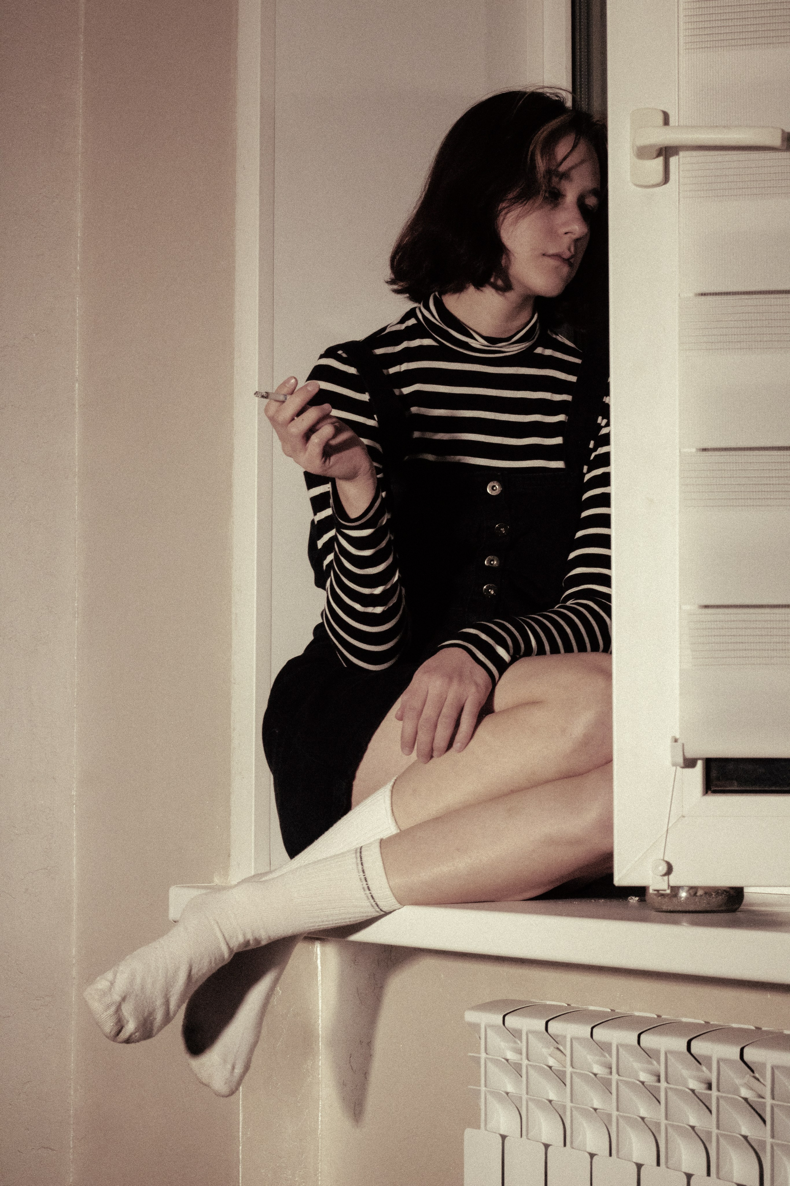 Девушка в полосатом свитере сидит на подоконнике, с сигаретой в руке, уставившись в пол — первое мгновение одиночества.