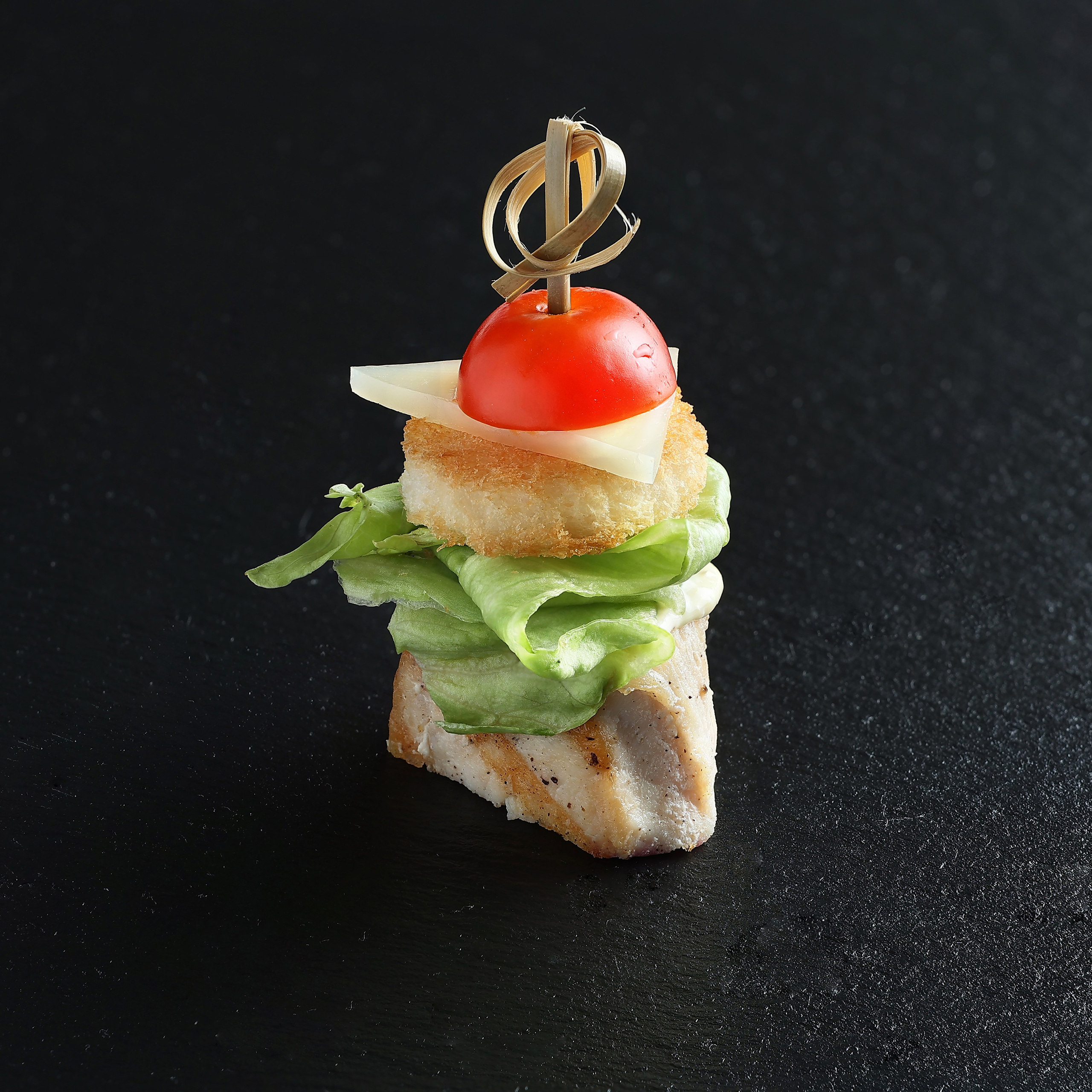Мини-закуска с курицей, салатом, сухариком, сыром и помидором черри на шпажке, на тёмном фоне.