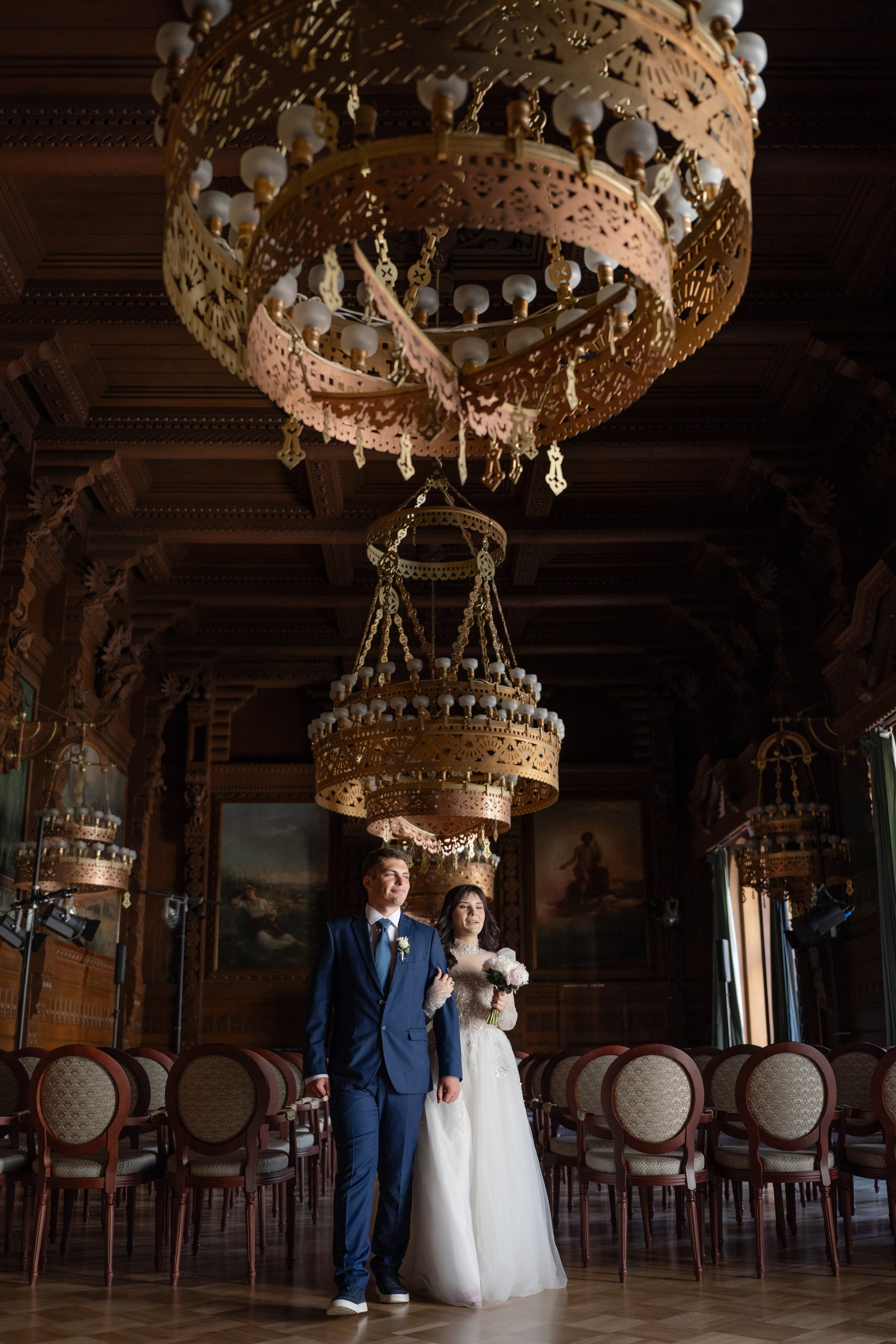 Свадебная фотосессия в Доме ученых, Дворце князя Владимира в Санкт-Петербурге. Жених с невестой в дубовом зале идут за руки под огромными люстрами