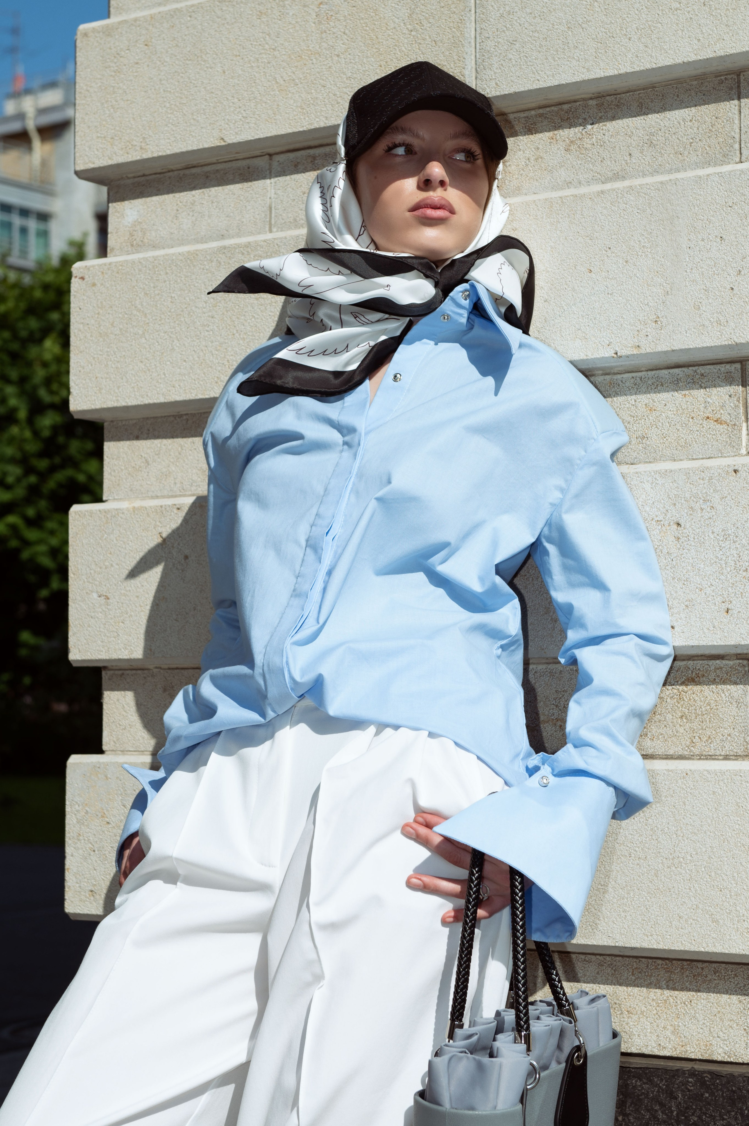 Красивая девушка в голубой рубашке и белых брюках и шелковом дизайнерском платке, повязанном поверх кепки стоит у стены