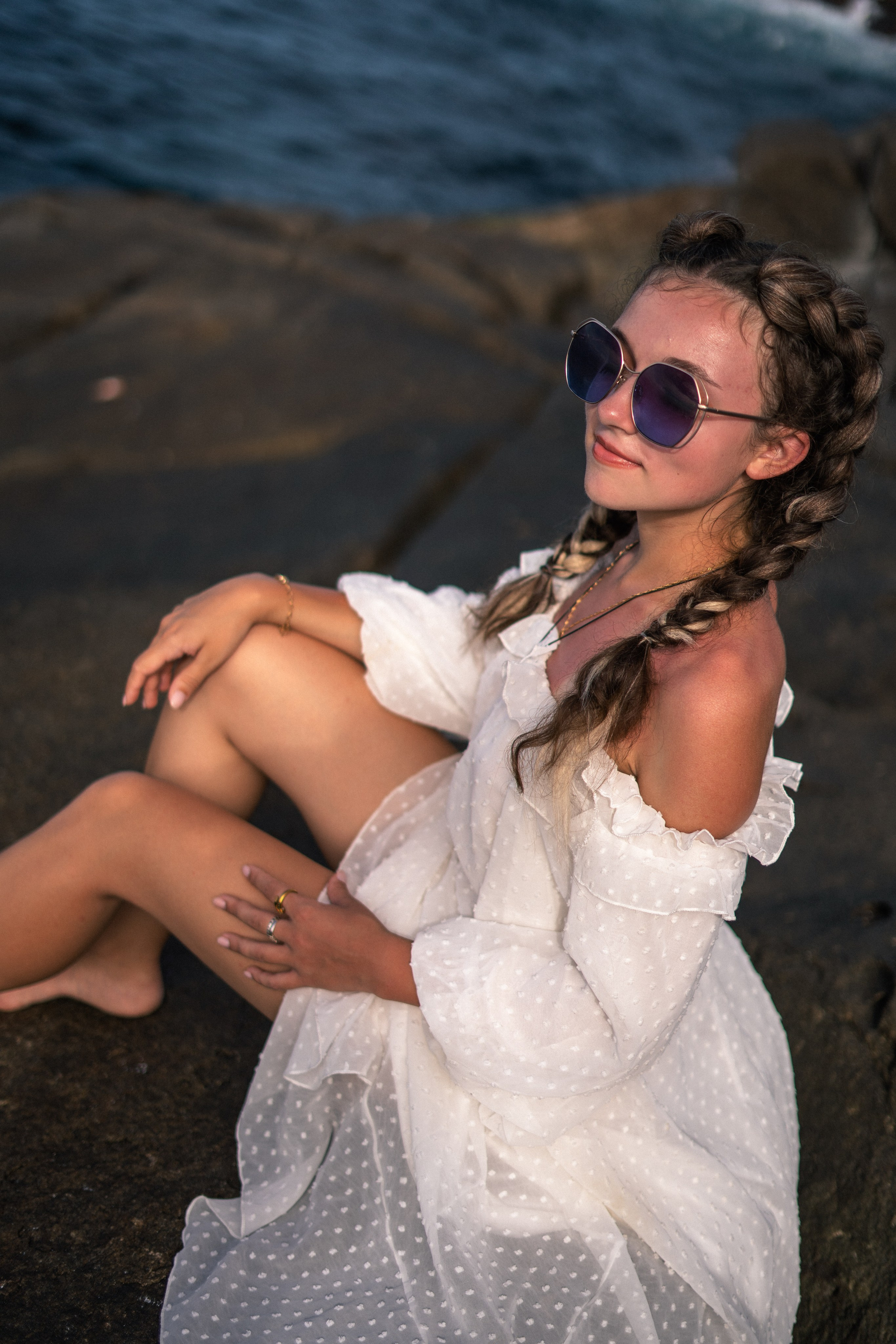 a young girl in a white dress and glasses laughing on the rocks by the ocean