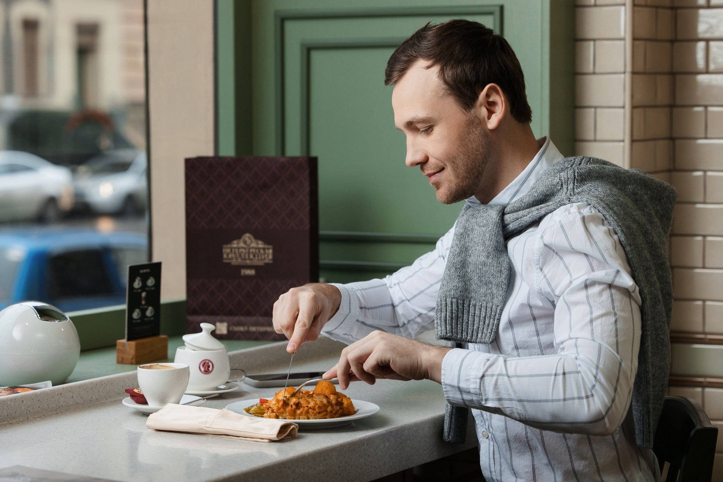 Парень с короткой щетиной в рубашке есть в ресторане у окна