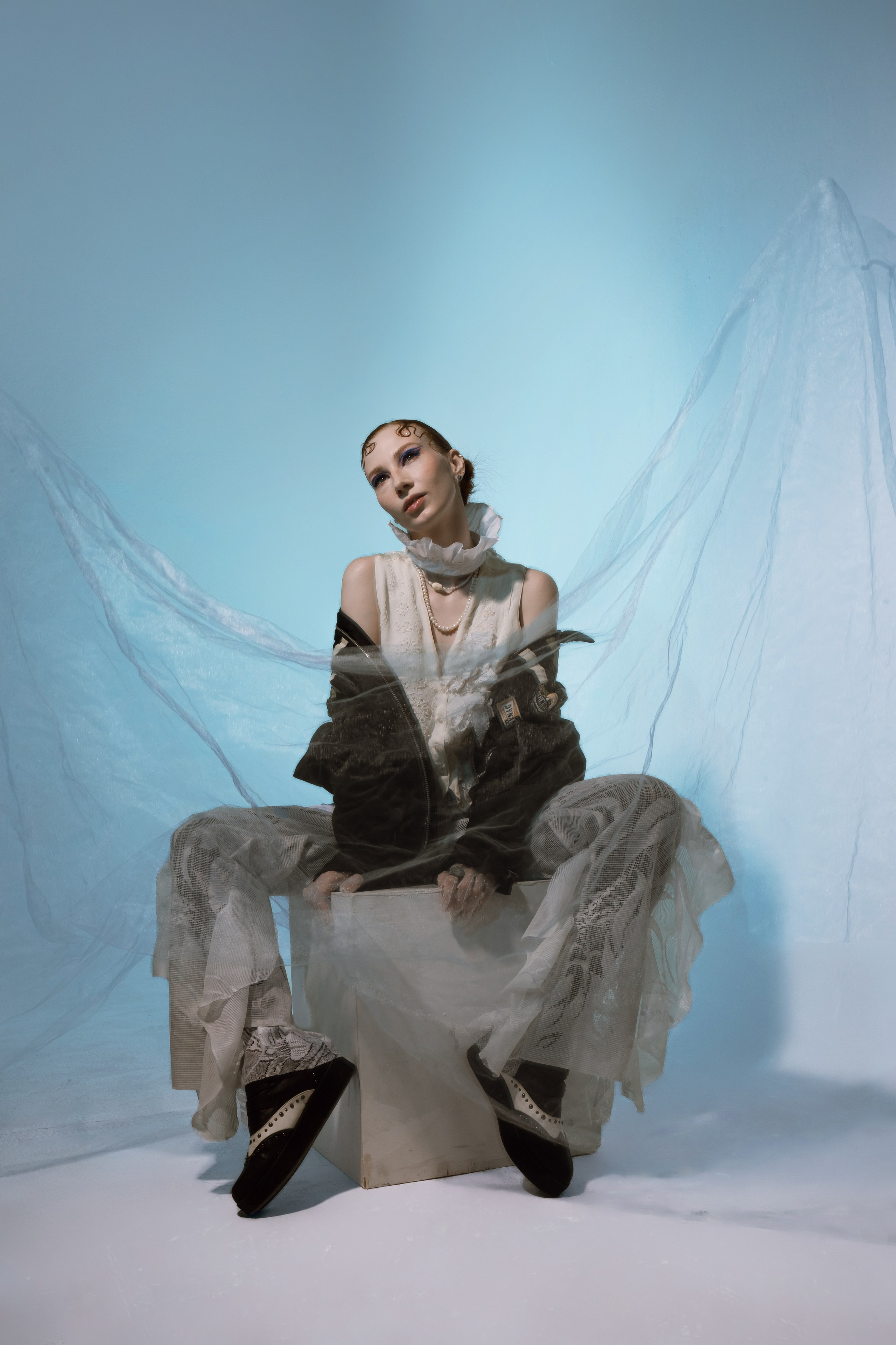 Модель представляет собой эффектный образ, украшенный замысловатыми тканями и аксессуарами, изящно сидящий на пастельно-голубом фоне с мягкой драпировкой.