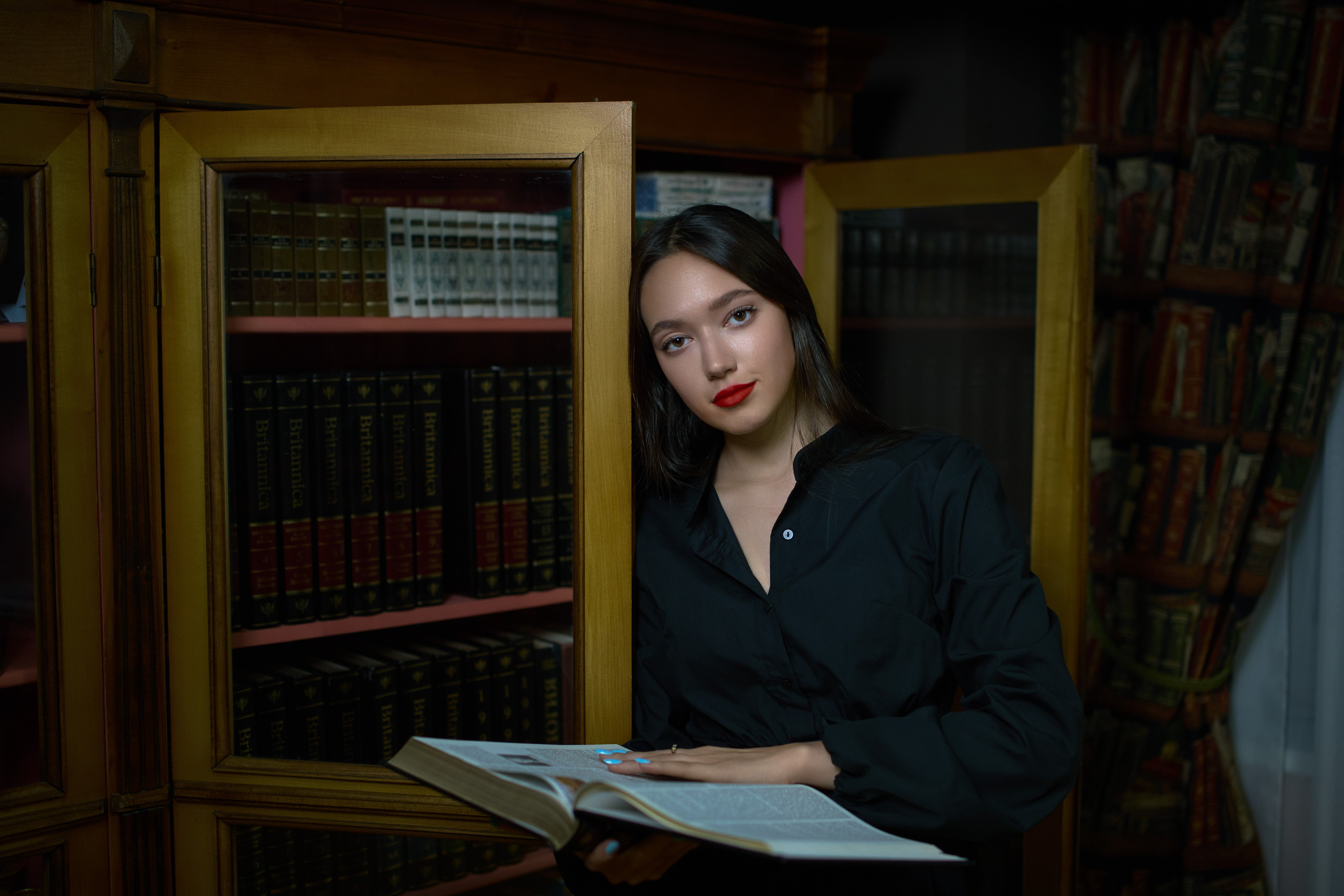 Девушка в черном платье стоит в библиотеке с раскрытой книгой у открытого шкафа