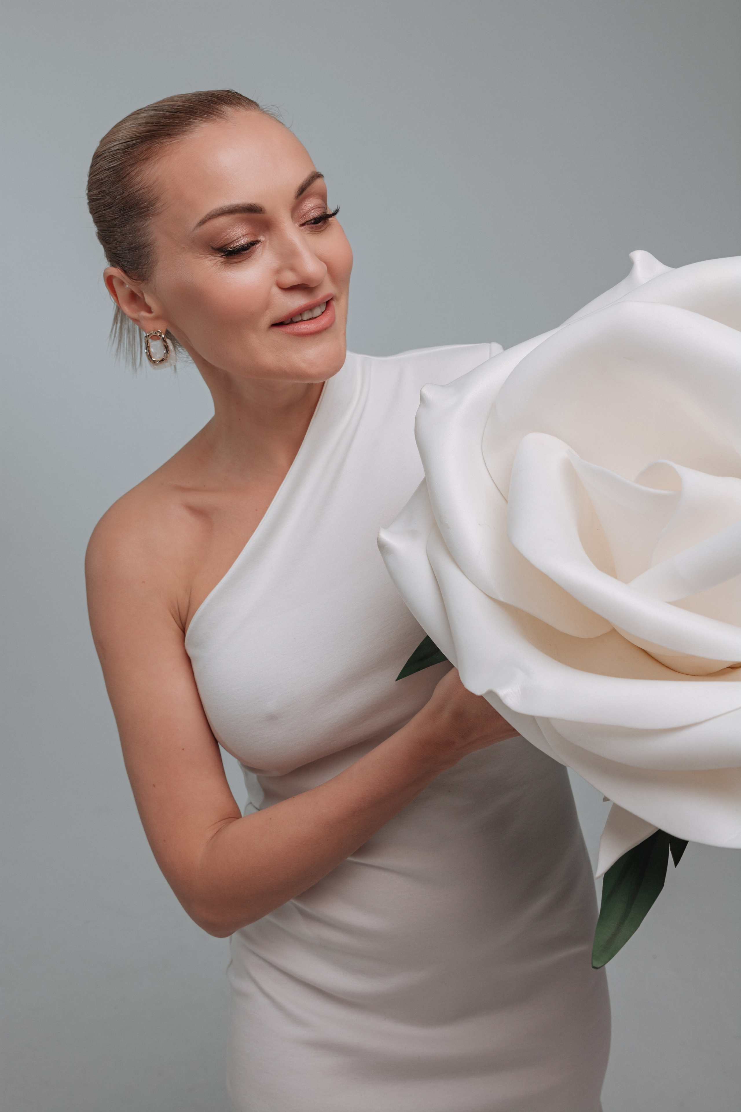 Женщина держит в руках белый цветок, возможно, лилию или другой крупный цветок с длинными лепестками. Цветок свежий и распустившийся, его белизна контрастирует с ее темными волосами, собранными в аккуратную прическу.