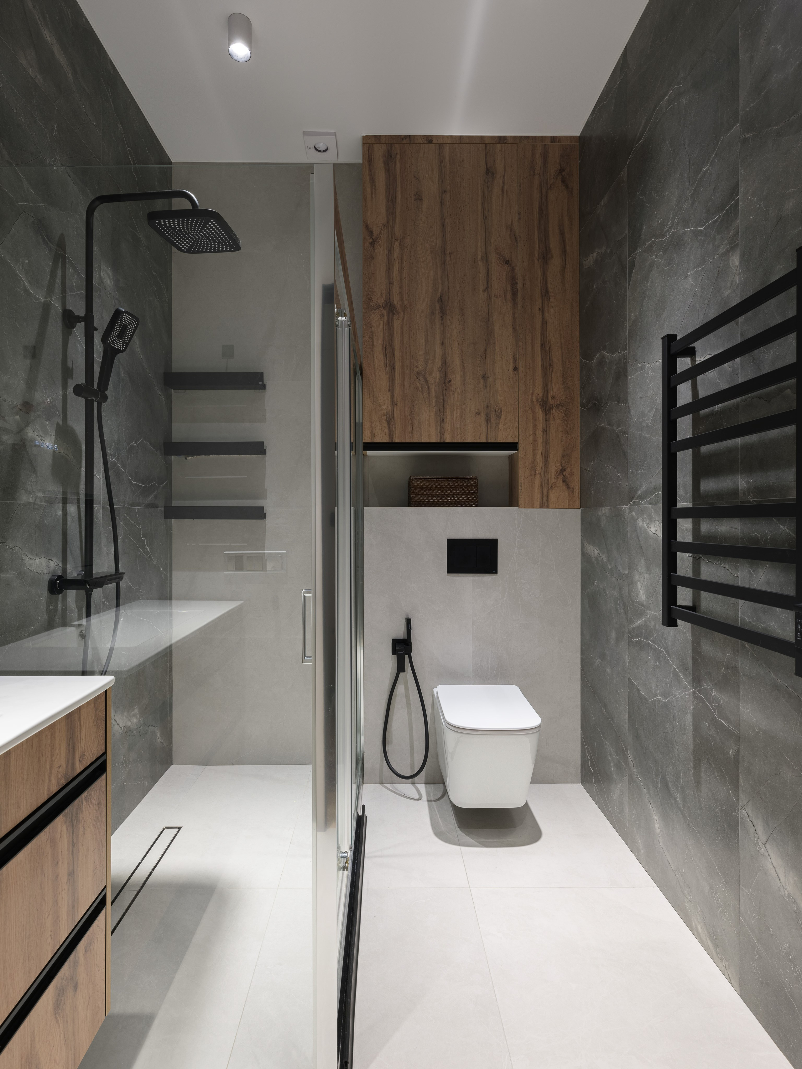 Современная ванная комната в минималистичном стиле с душевой кабиной из стекла, подвесным унитазом и плиткой под серый камень. Контрастная черная сантехника и деревянные панели создают уют и динамику пространства. Дизайн Алина Дёрова, стилист Наталья Якушева, фото Сергей Денисенко.