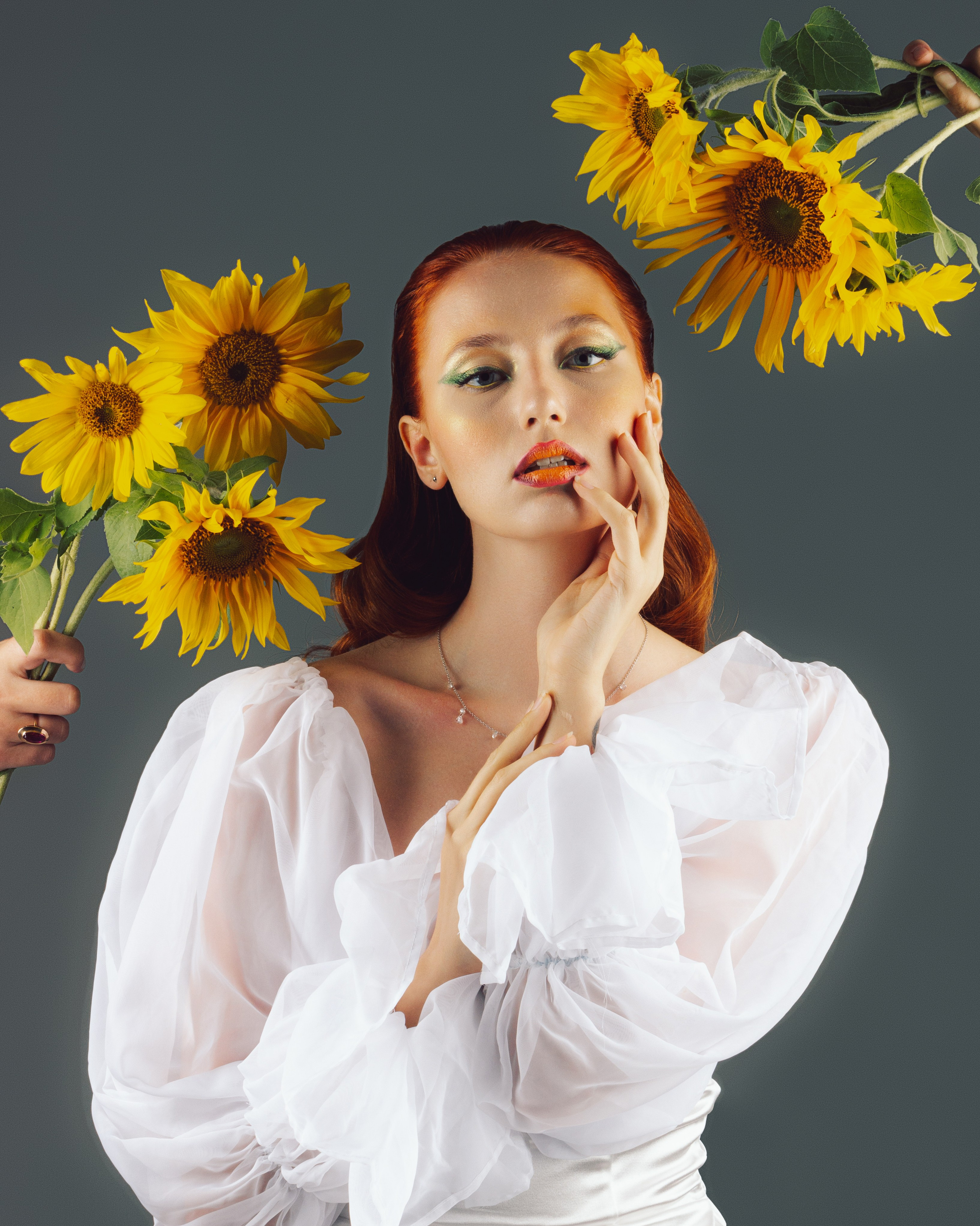 крупный портрет девушки, за ее спиной расположены несколько букетов подсолнухов на сером фоне 