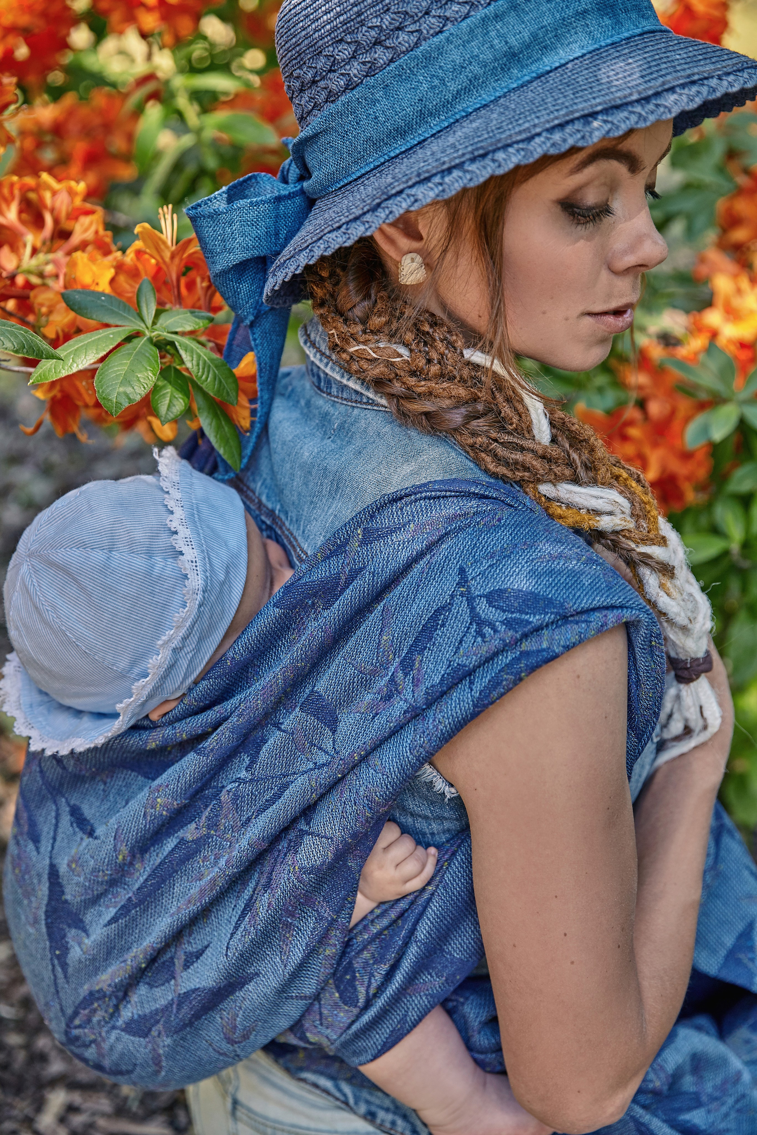Синий слинг, малыш за спиной у мамы на фоне оранжевых цветов