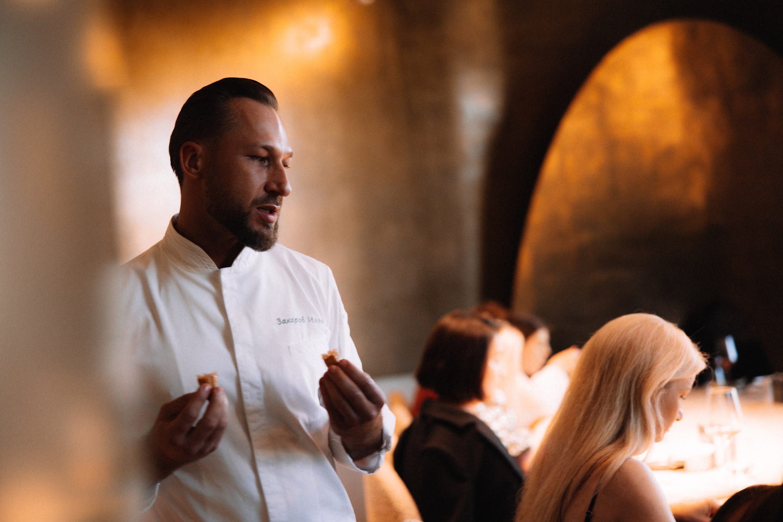 Мероприятие: Шеф-повар Илья Захаров в белом кителе рассказывает о блюдах гостям вечера.