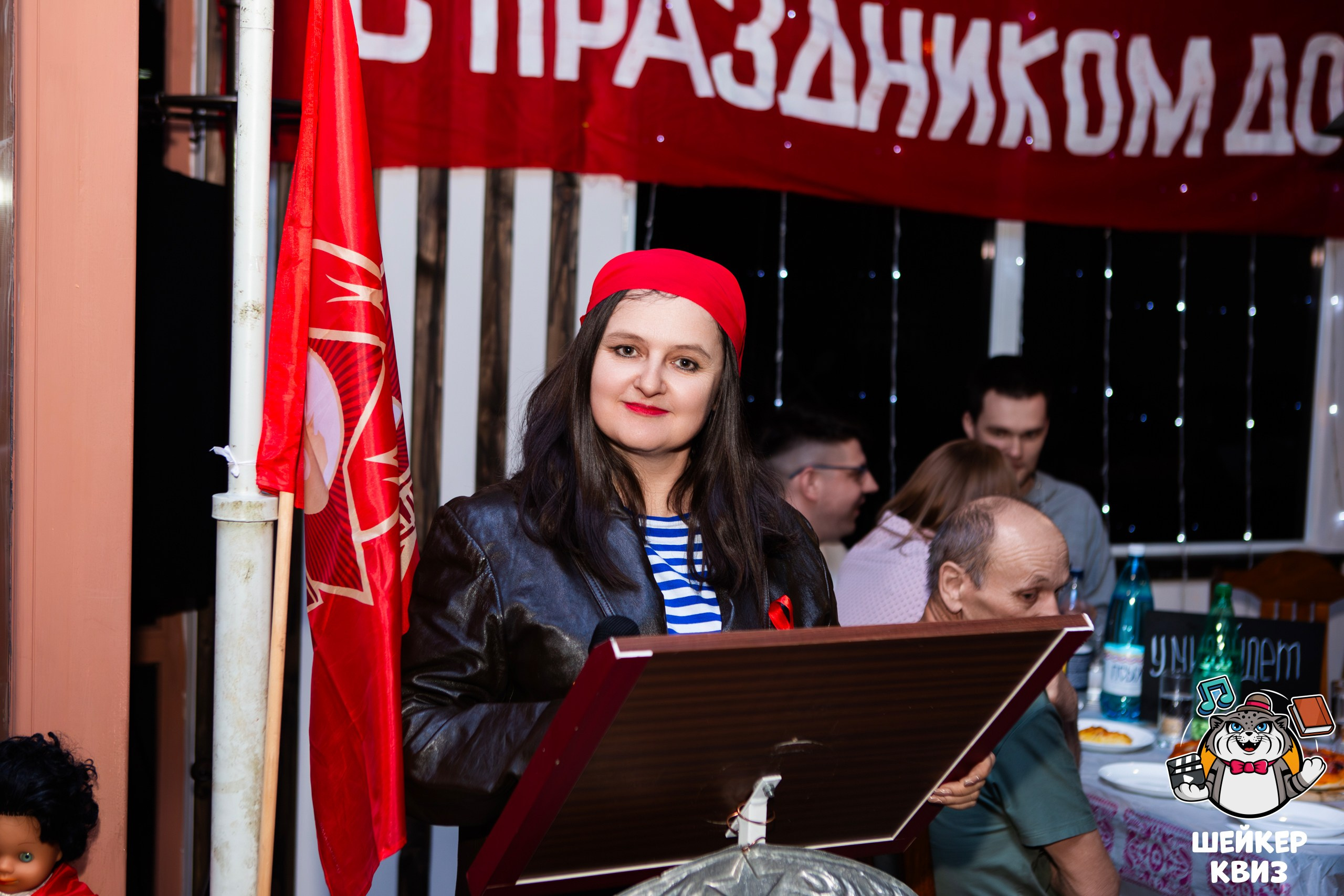 Женщина в красном берете и полосатой рубашке стоит на трибуне с красным флагом в Туле, а люди сидят за столами позади неё; над ними висит праздничное знамя. Профессиональные фотографии с корпоративов от фотографа Николая Богатырева.