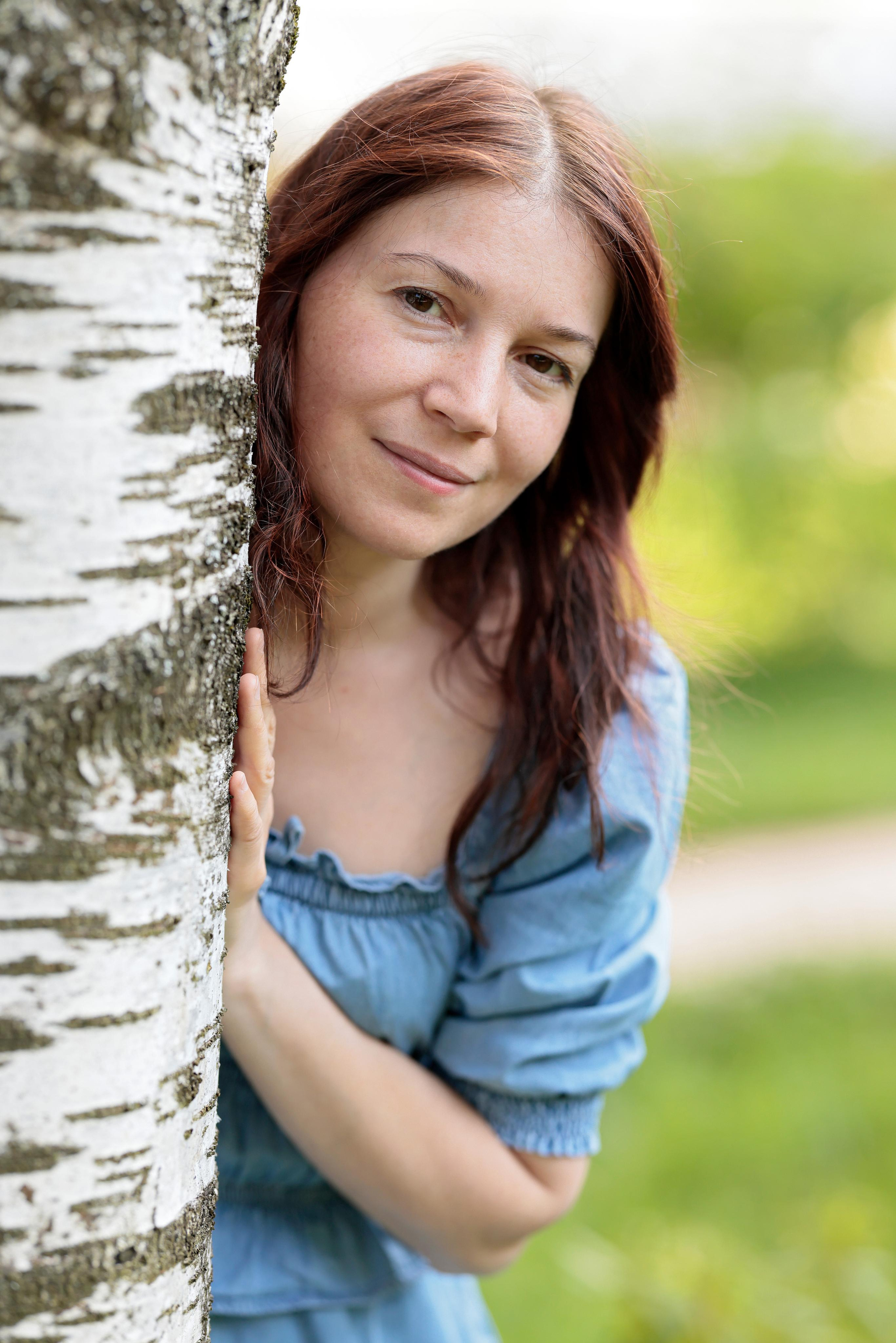 Портрет женщины в голубом платье, выглядывающая из-за ствола берёзы на фоне зелени.