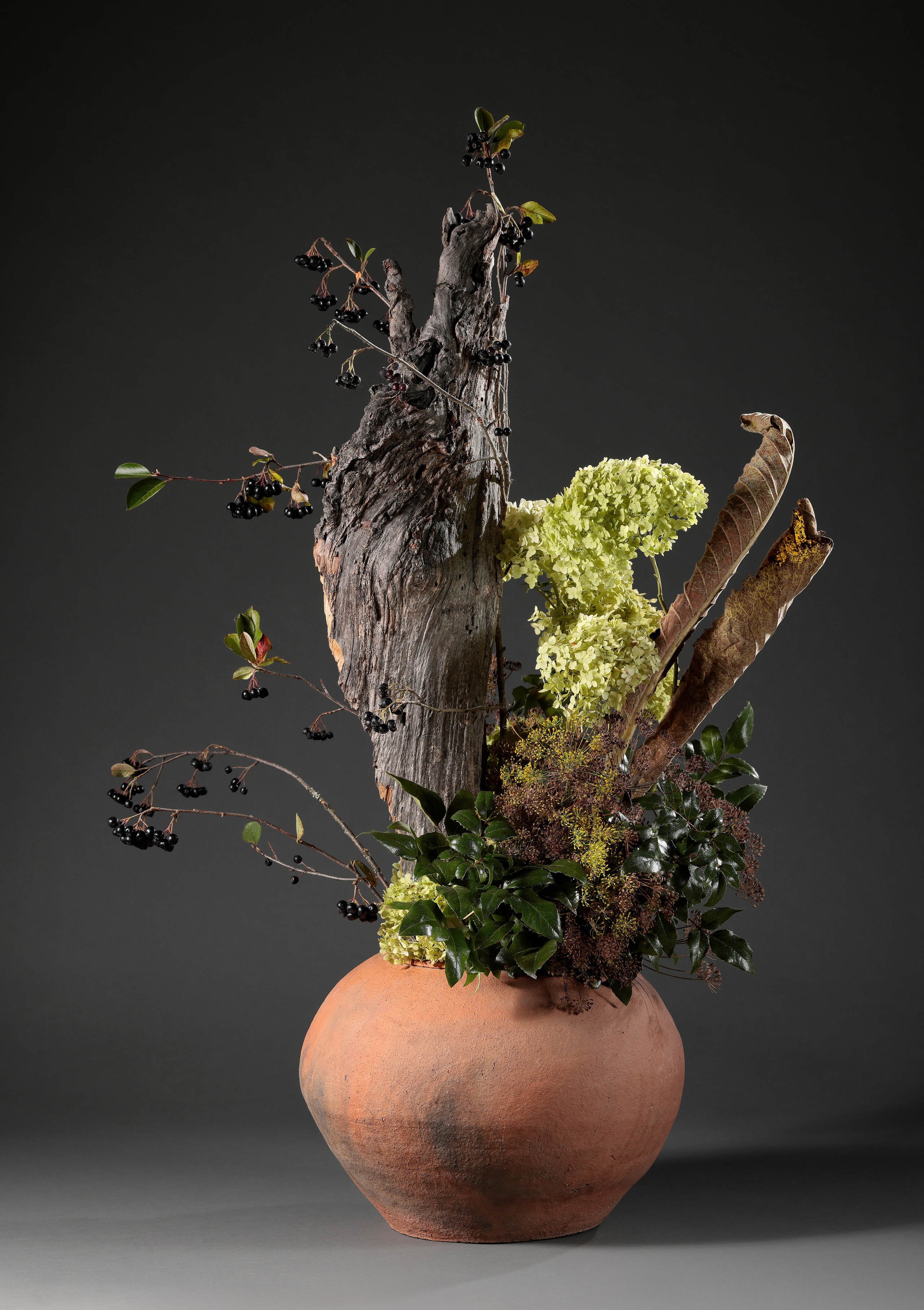 Икебана в глиняной круглой вазе с корой и ветками на сером фоне.
