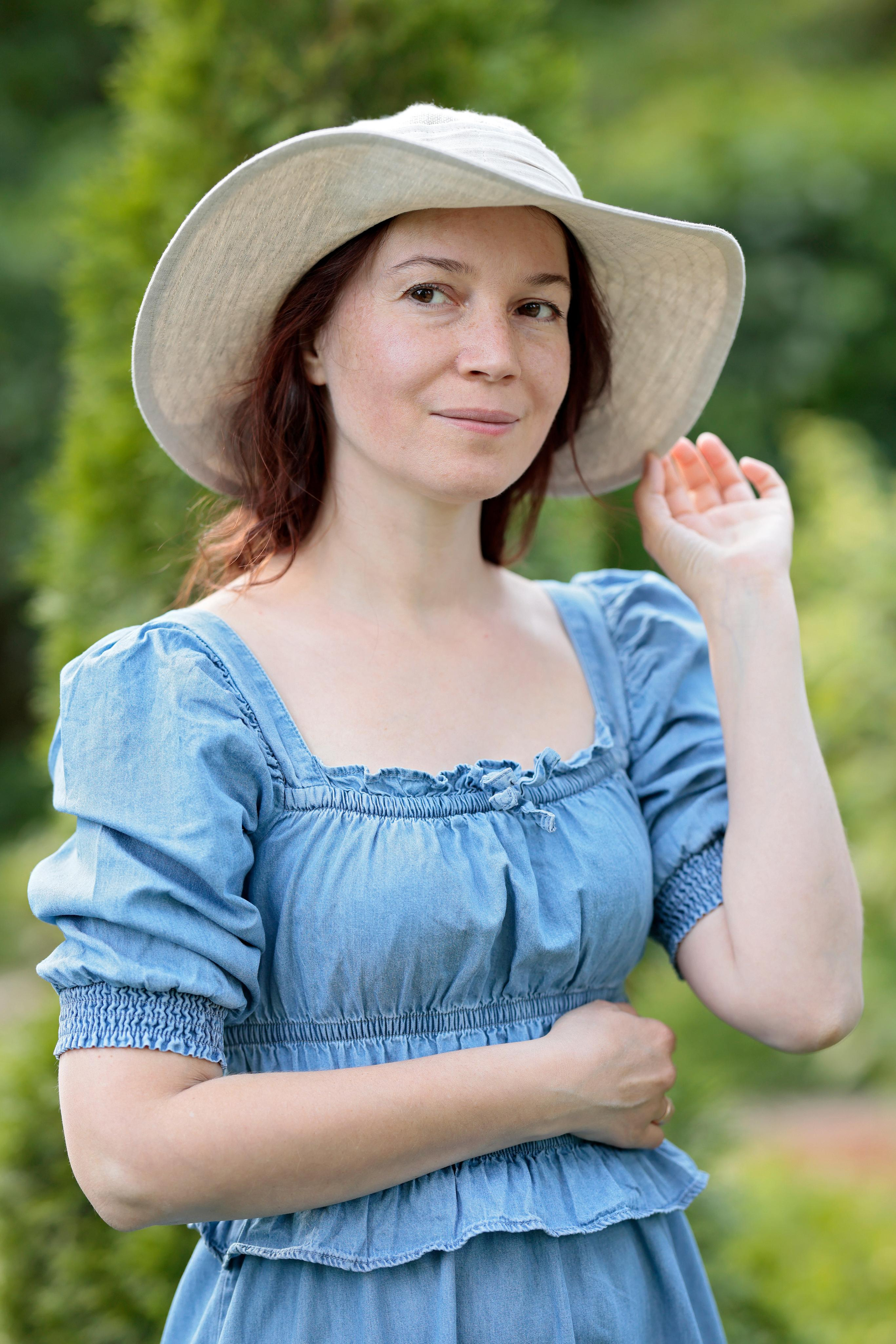 Портрет женщины в шляпе и голубом платье на фоне зелени.