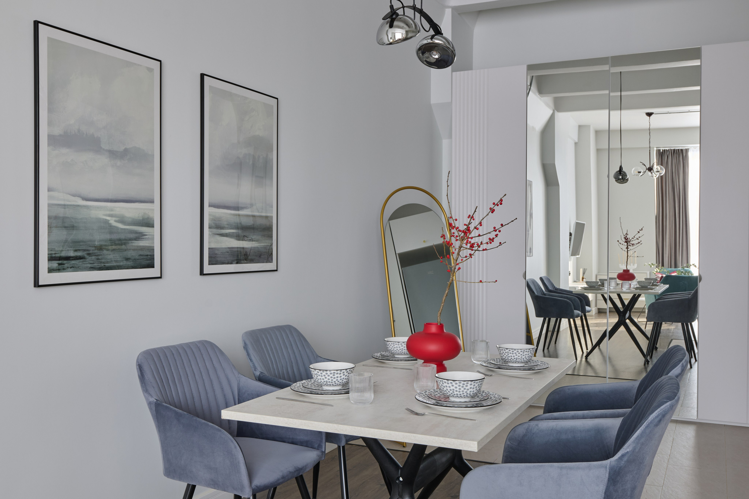 Обеденная зона с синими велюровыми стульями вокруг светлого обеденного стола. На стене висят две картины с морскими пейзажами, а на столе красная ваза с ветками.