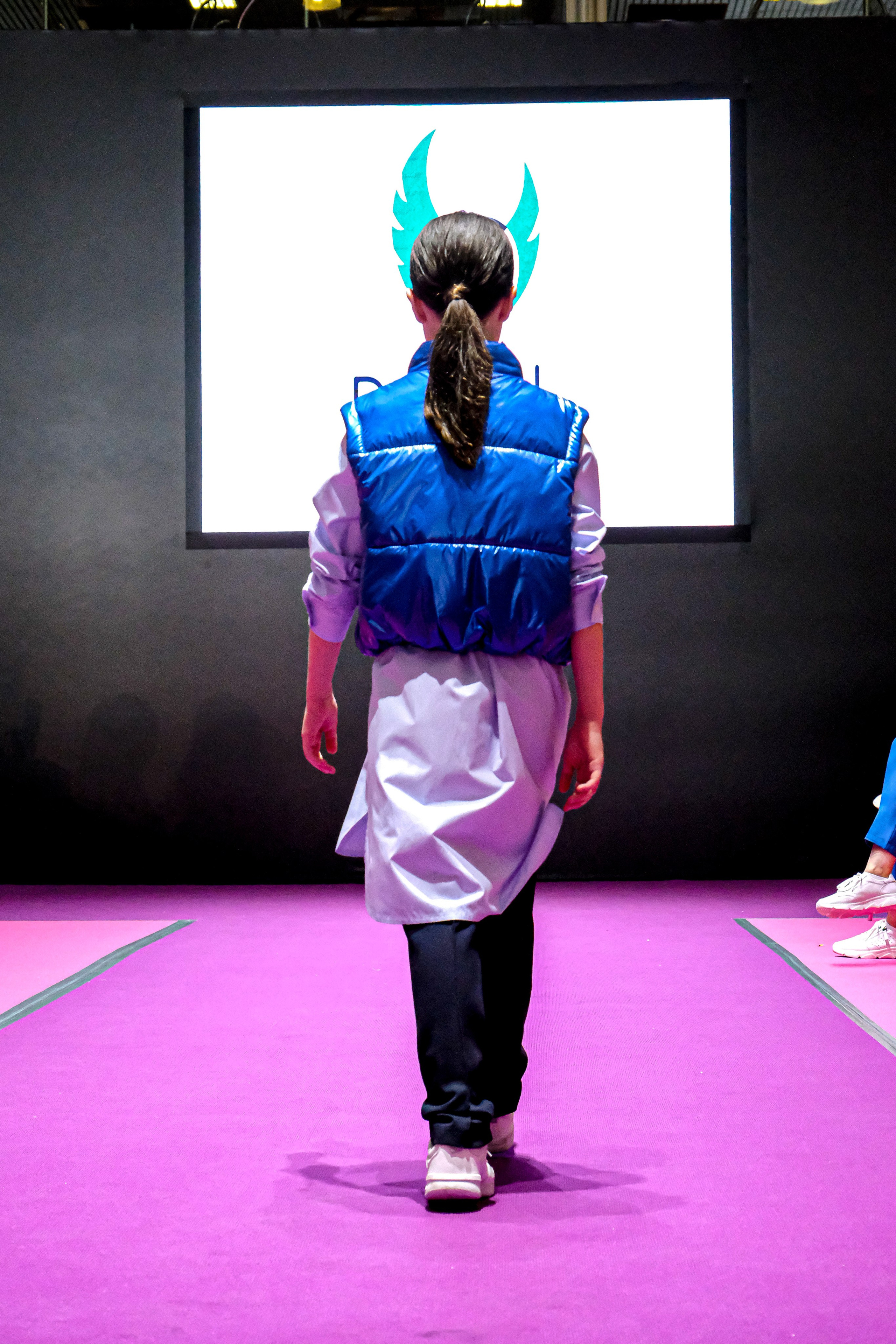  "Модели в ярких нарядах проходят по подиуму во время показа мод, освещенные яркими софитами и окруженные восторженными зрителями."