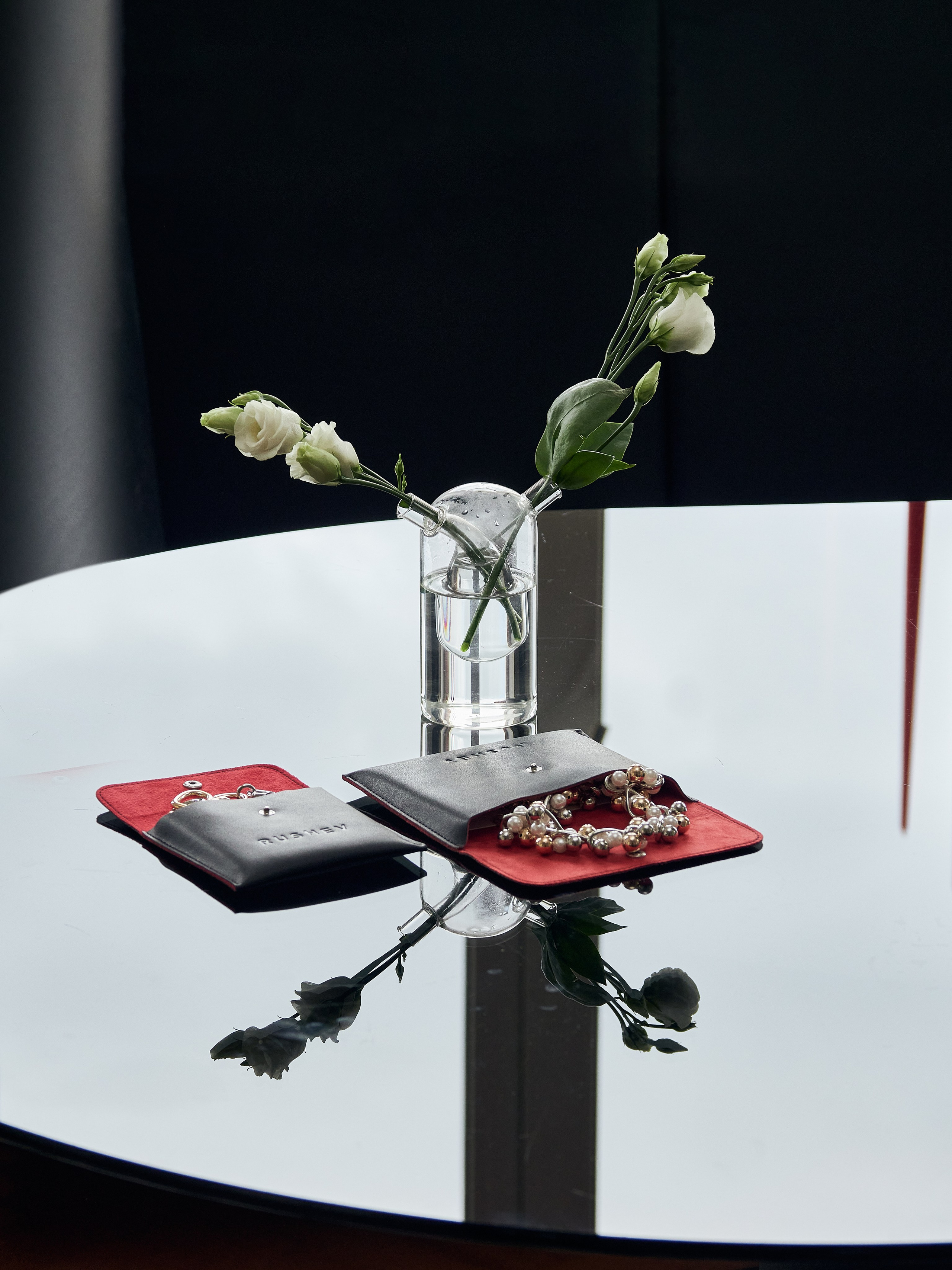 Стеклянная ваза с белыми цветами стоит на отражающем круглый стеклянный стол. Рядом с ним лежат черно-красные бархатные мешочки для драгоценностей, в одном из которых находится серебряный браслет, а в другом — золотистый браслет с жемчужными вставками.