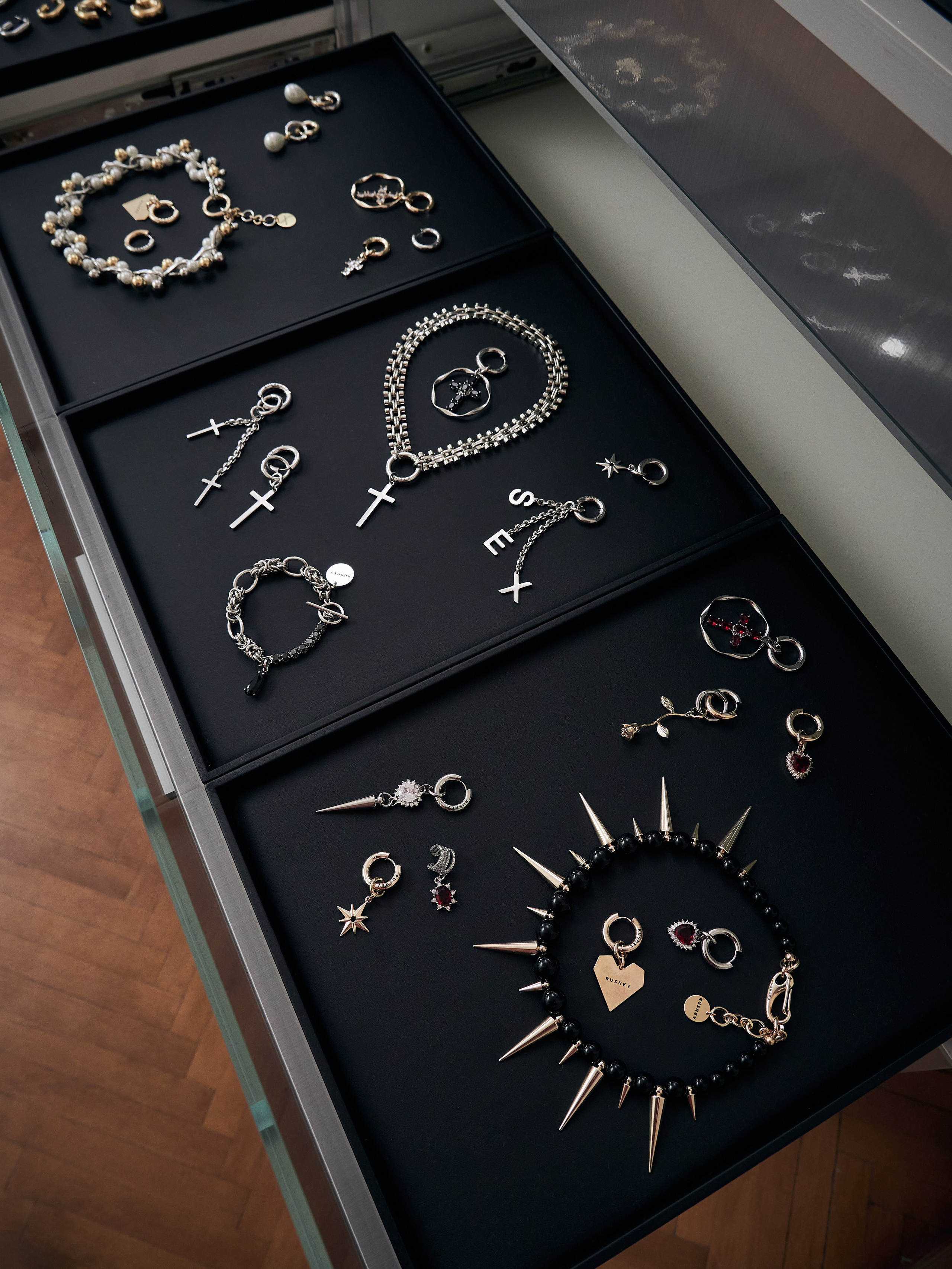 На выставке представлены различные ювелирные изделия, в том числе ожерелья, кольца, серьги и браслеты. Некоторые украшены жемчугом, крестами, шипами и сердечками. Предметы элегантно разложены на черных подносах внутри стеклянной витрины на деревянном полу.