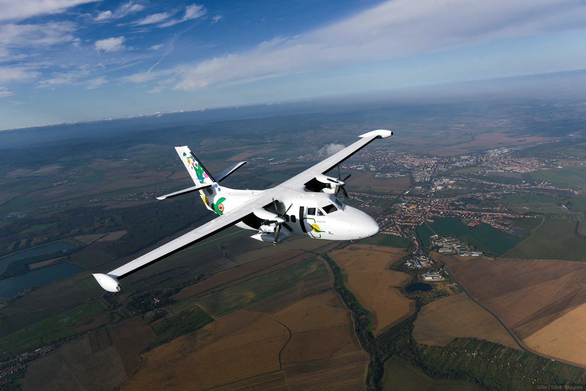 Двухдвигательный турбовинтовой самолет Let L-410 Turbolet в полете над равнинной местностью с населенными пунктами. Воздушное судно белого цвета с рисунками на фюзеляже и хвосте. Фото: Гелио.