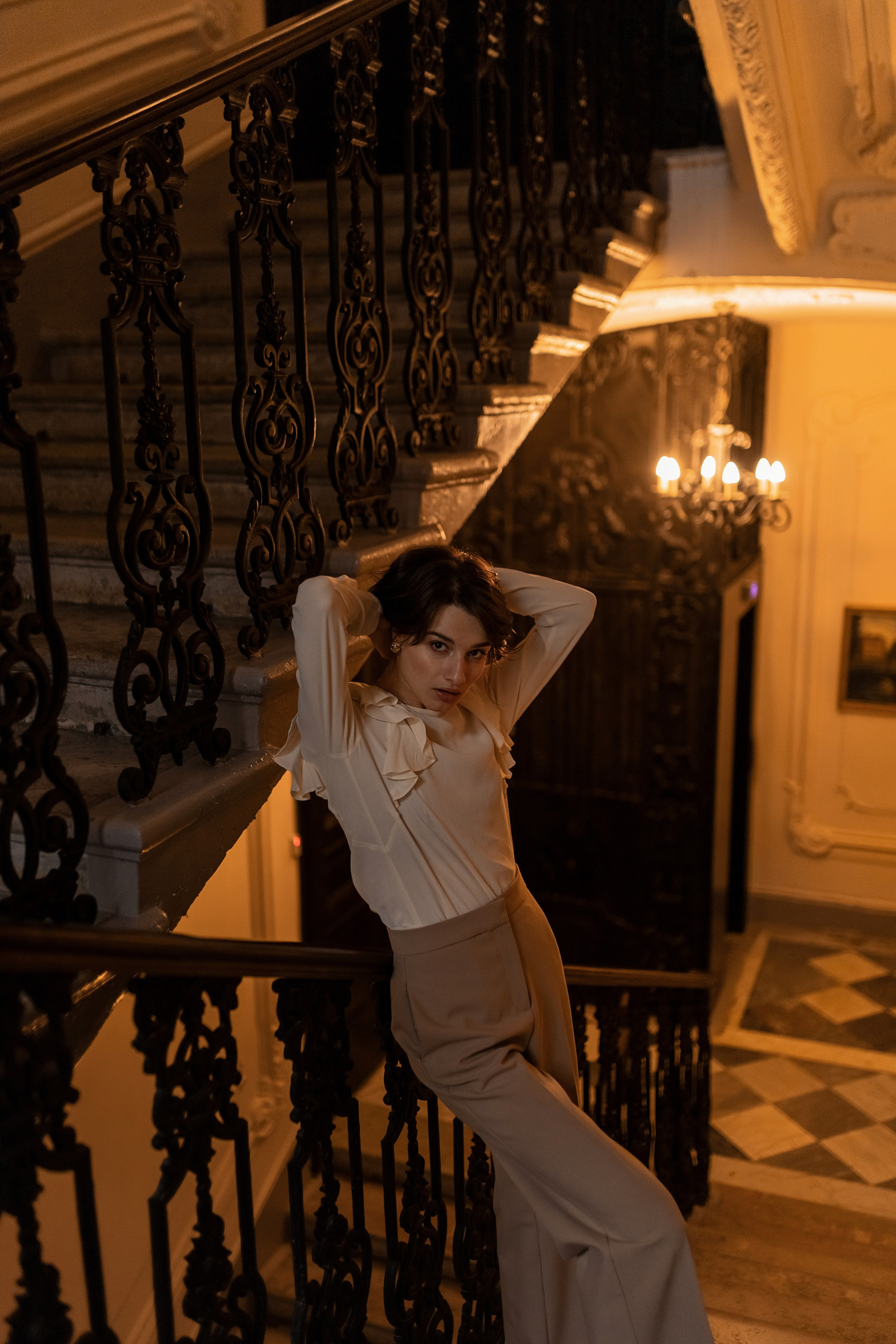 При теплом освещении женщина принимает шикарную позу на богато украшенной лестнице парадной, демонстрируя свой изысканный наряд и красоту помещения.