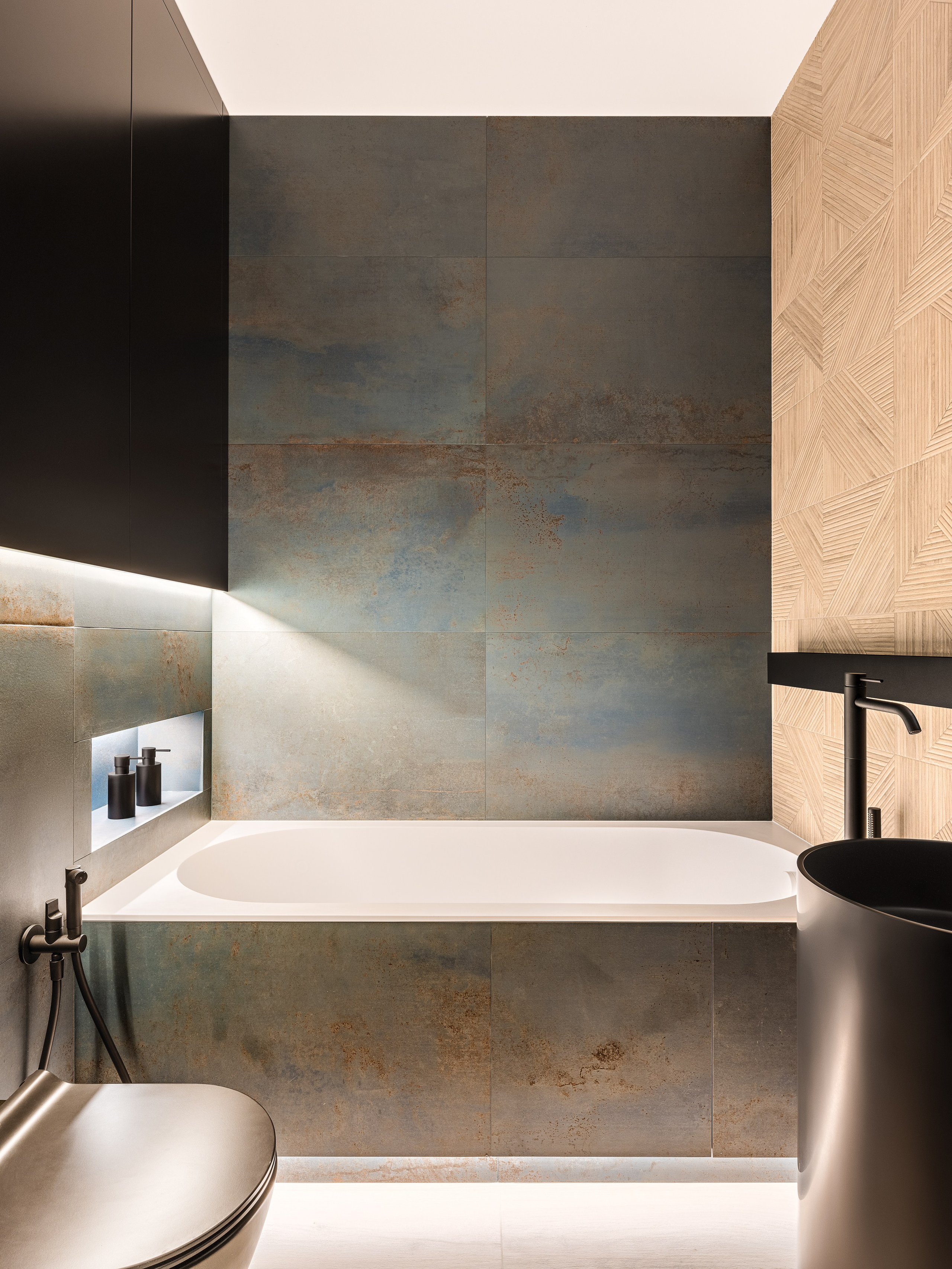Интерьер ванной комнаты с прямоугольной встроенной ванной, серо-синими плитами с эффектом ржавчины, встроенной нишей с аксессуарами, черной сантехникой и комбинированной отделкой стен. Дизайнеры Ренат Байчуков и Алеся Кириенко, стилист Екатерина Ракитина, фотограф Сергей Денисенко.
