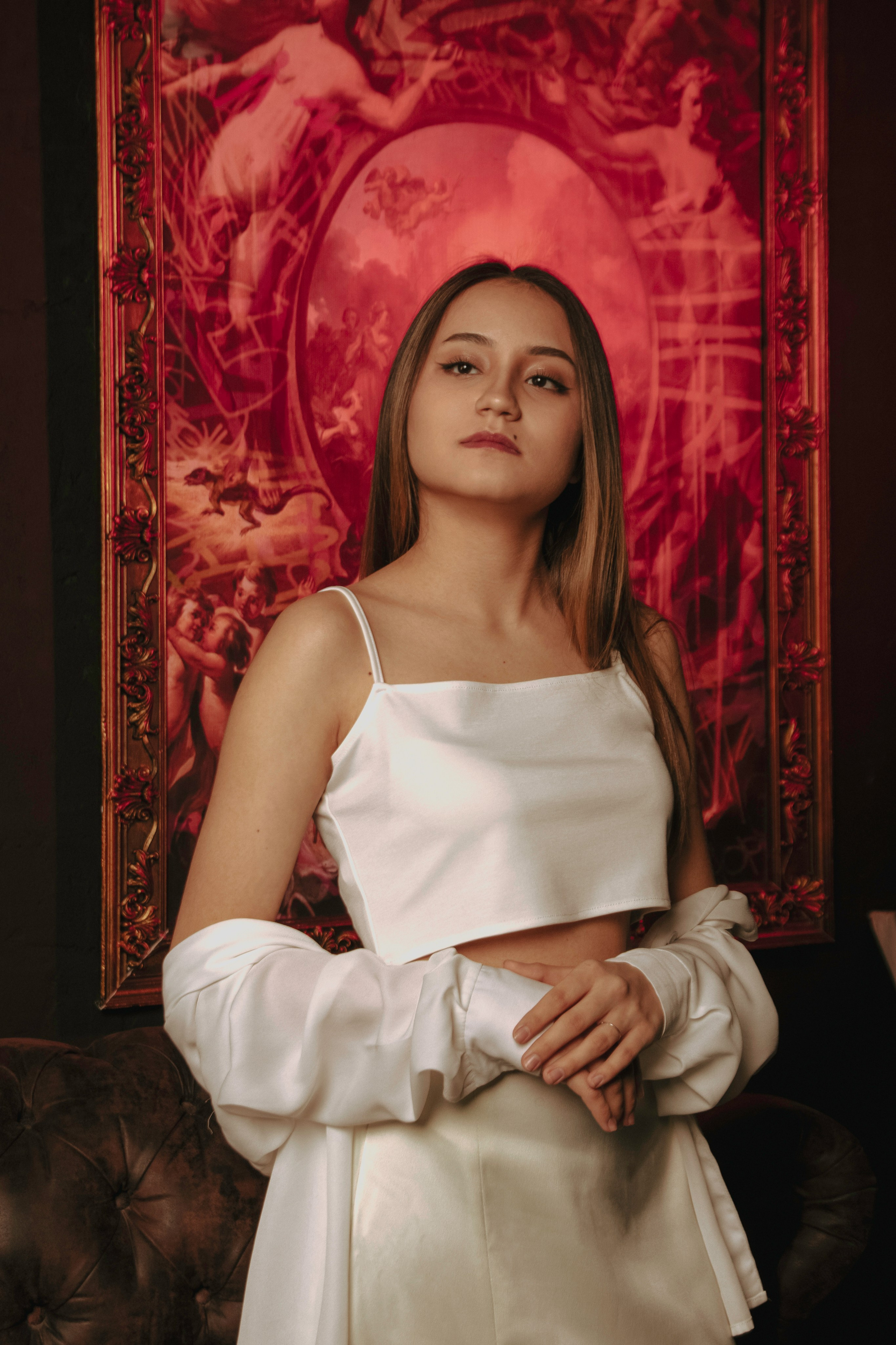 Девушка в стиле ренессанс в белой одежде фотографируется на фоне подсвеченной красным картины 