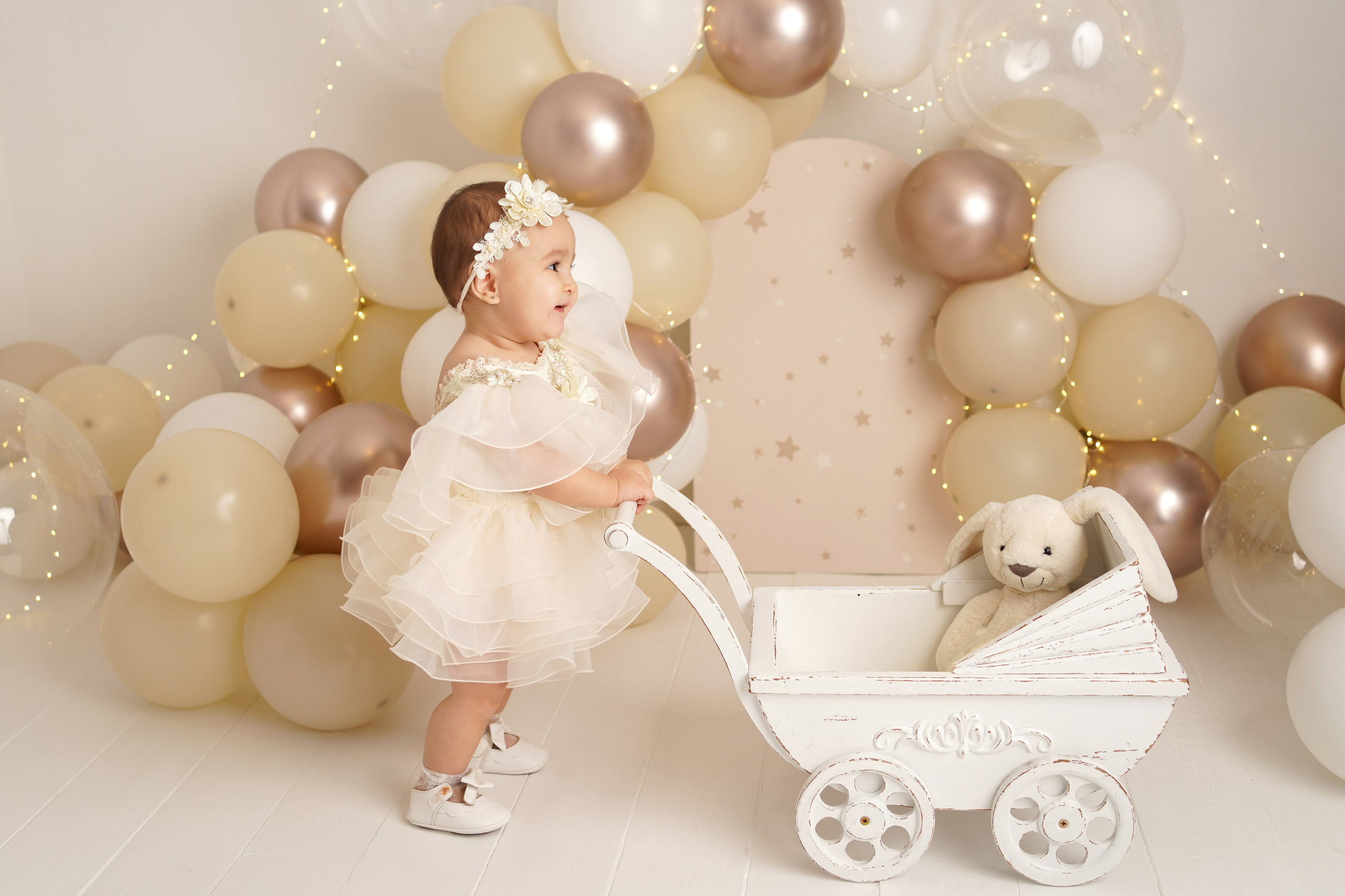 Малышка в бежевом  платье и цветочном веночке катает детскую коляску с зайкой.