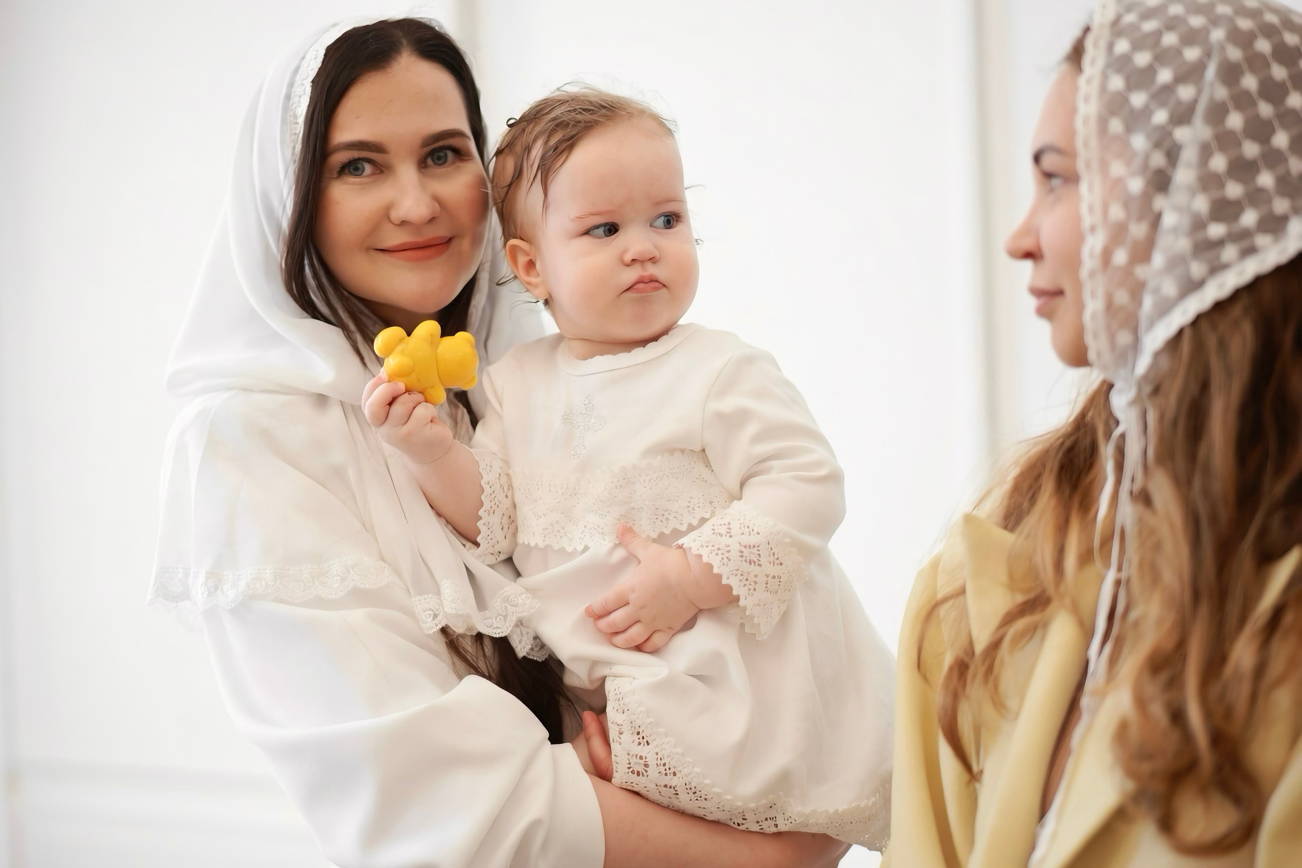 Крестная мать держит младенца, пожилой священник на втором плане, мягкий белый свет — Екатеринбург, Илья Антропов