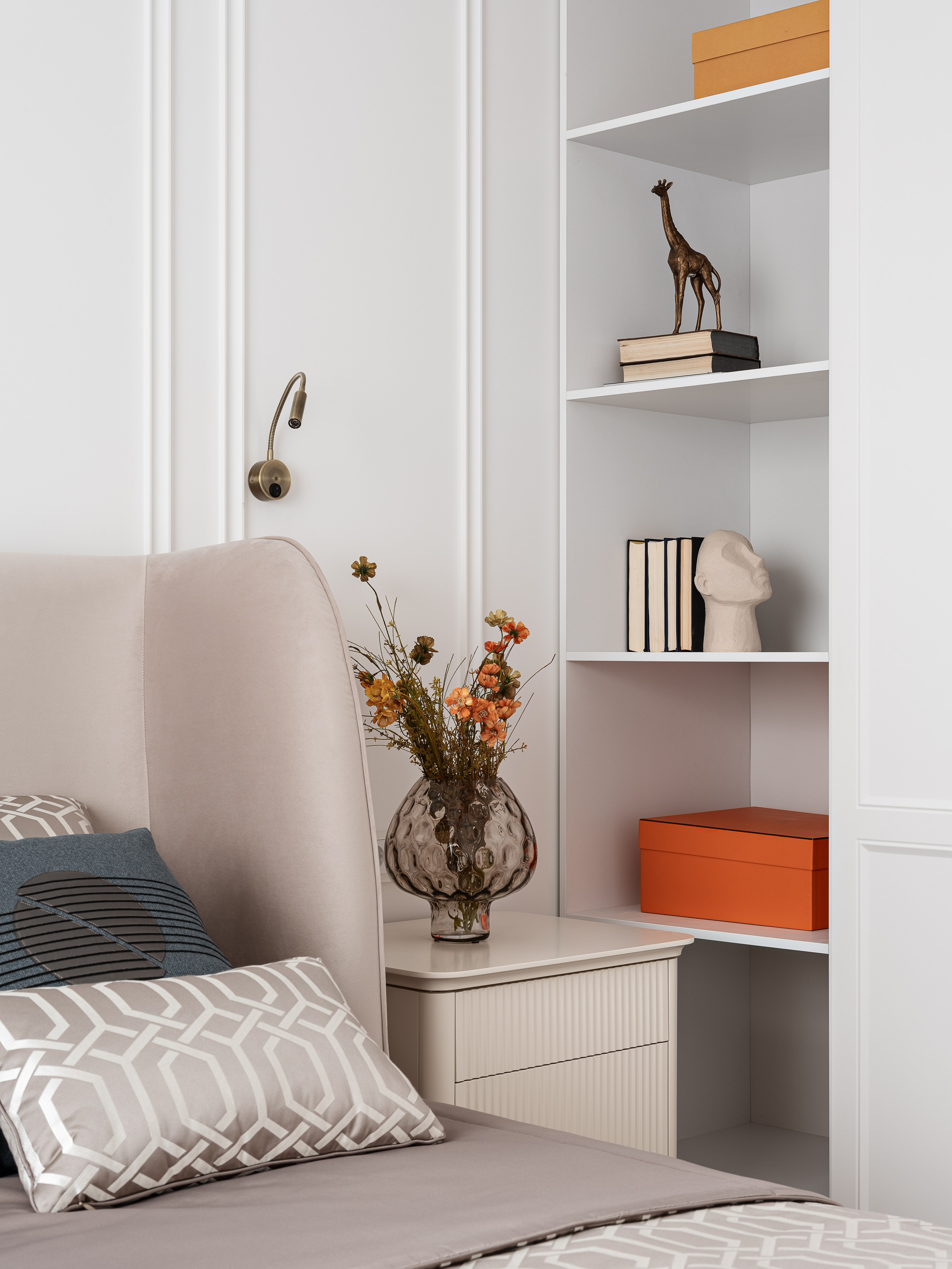 Элегантный интерьер спальни с встроенными белыми полками, декоративными книгами, фигуркой жирафа, скульптурой, вазой с цветами и яркой коробкой, оформленный дизайнером Василиссой Бачевой, стилист Натальей Якушевой и снятый фотографом Сергеем Денисенко.