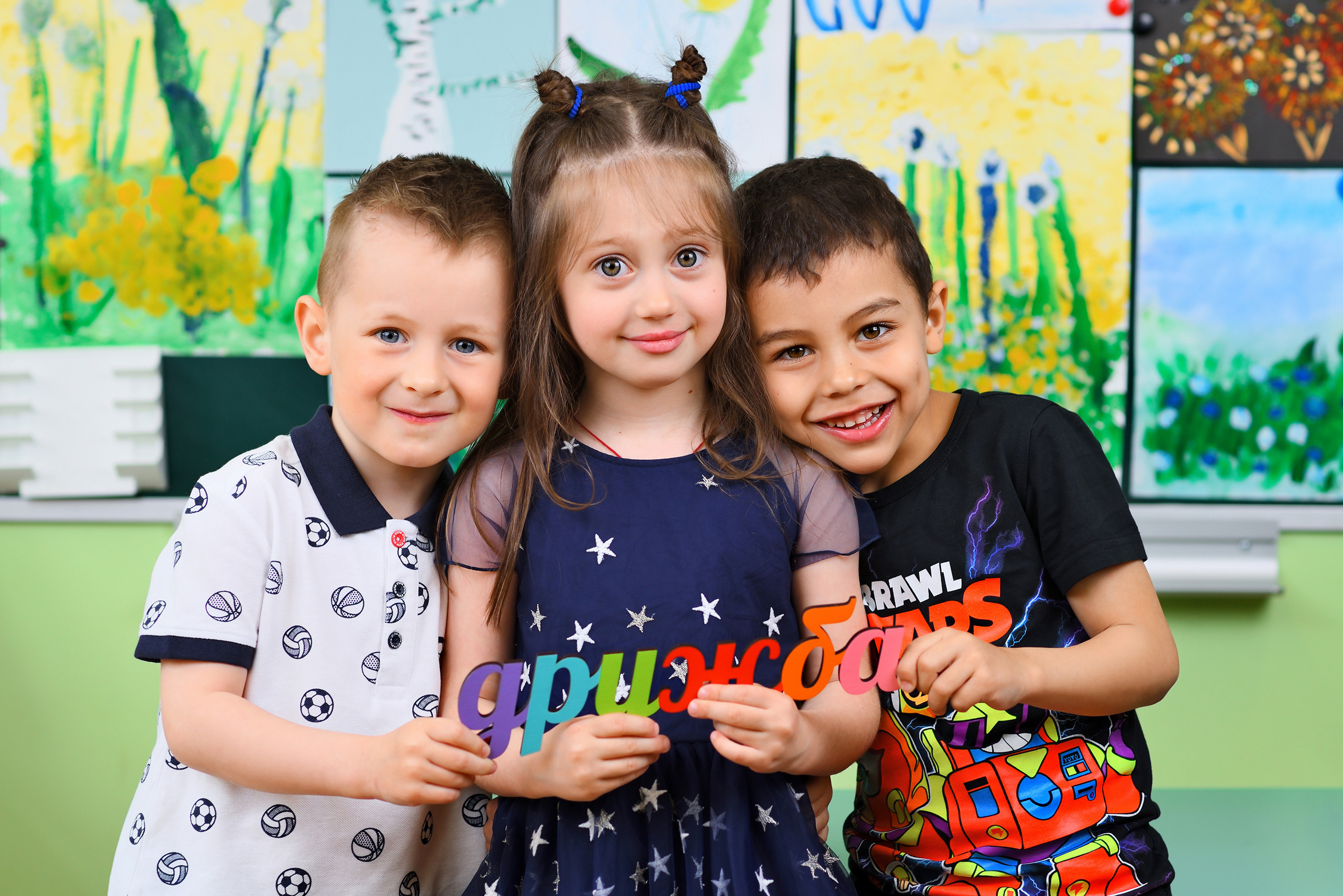 Дети в детском саду обнимаются с табличкой ДРУЖБА в руках.