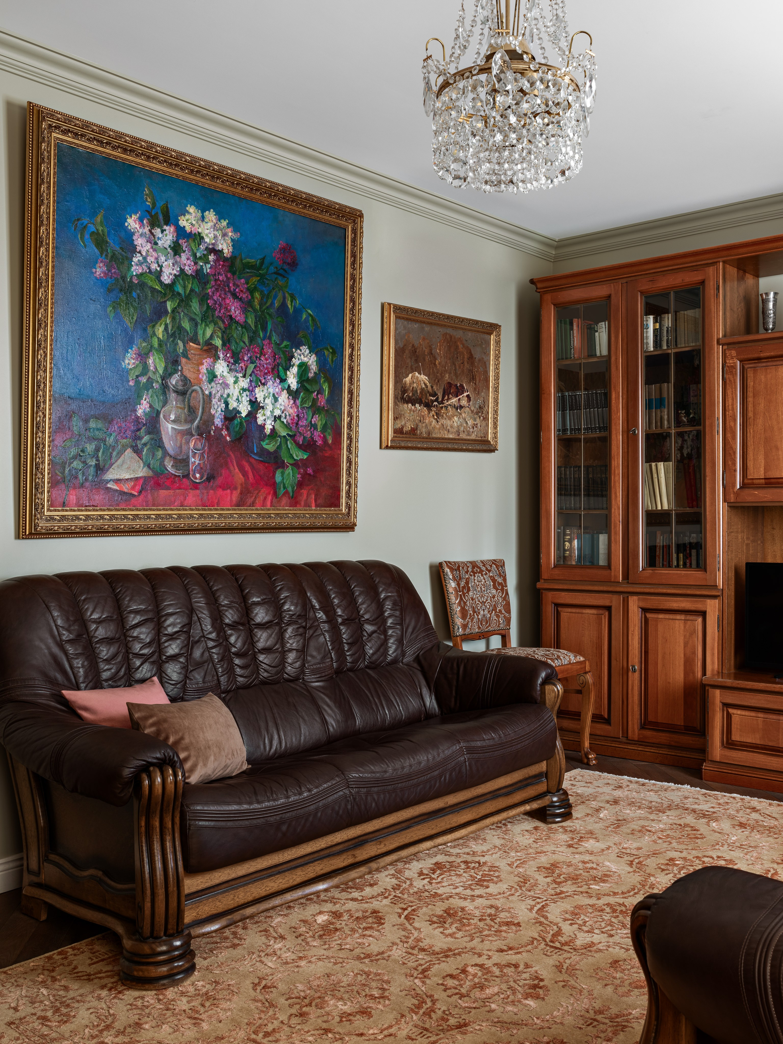 Интерьер гостиной с кожаным диваном, ковром с орнаментом и крупными живописными полотнами в золочёных рамах; хрустальная люстра добавляет изысканности. Проект дизайнеров Юлии Капункиной и Альбины Терещенко, стилист Алина Нелень, фото Сергея Денисенко.