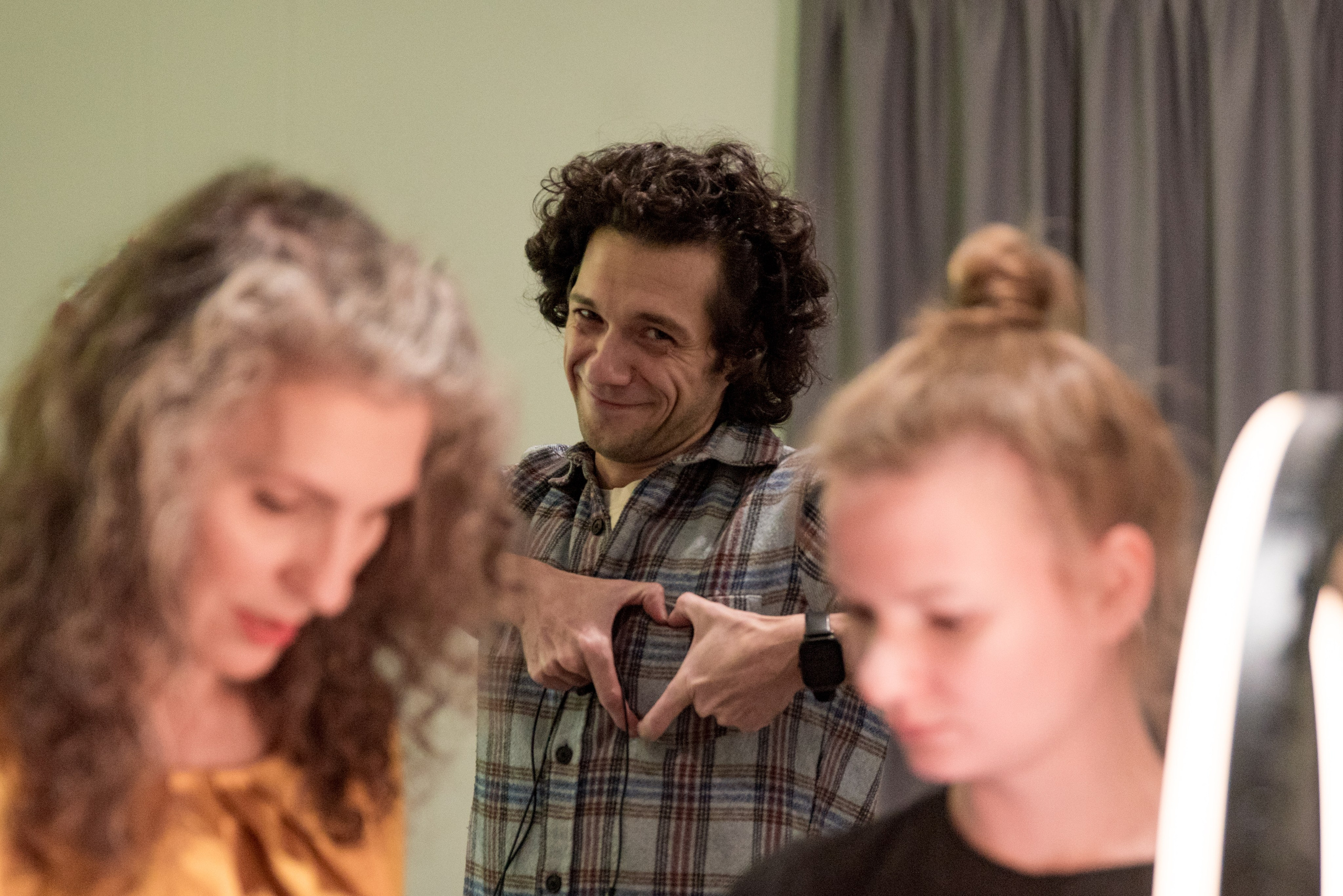 На переднем плане два размытых женских лица, что-то рассматривают. На заднем мужчина смотрит в камеру улыбается и показывает сердечко из пальцев.