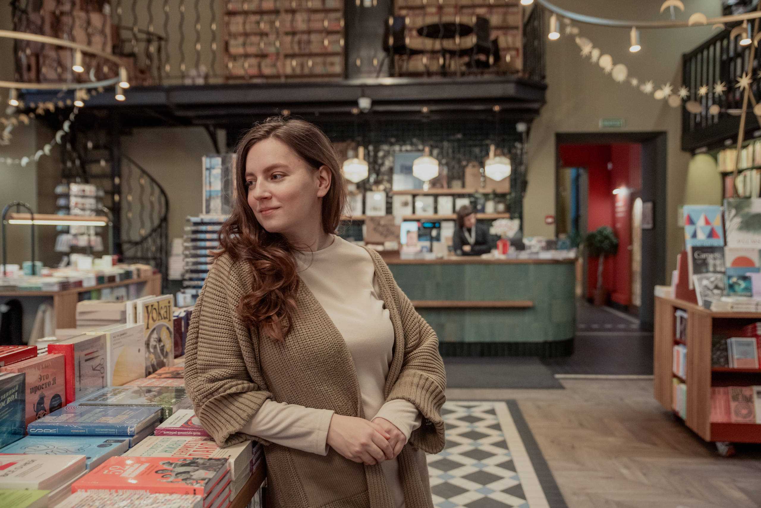 Большой книжный магазин. Женщина с длинными волосами в  бежевой кофте стоит облокотившись на прилавок с книгами.
