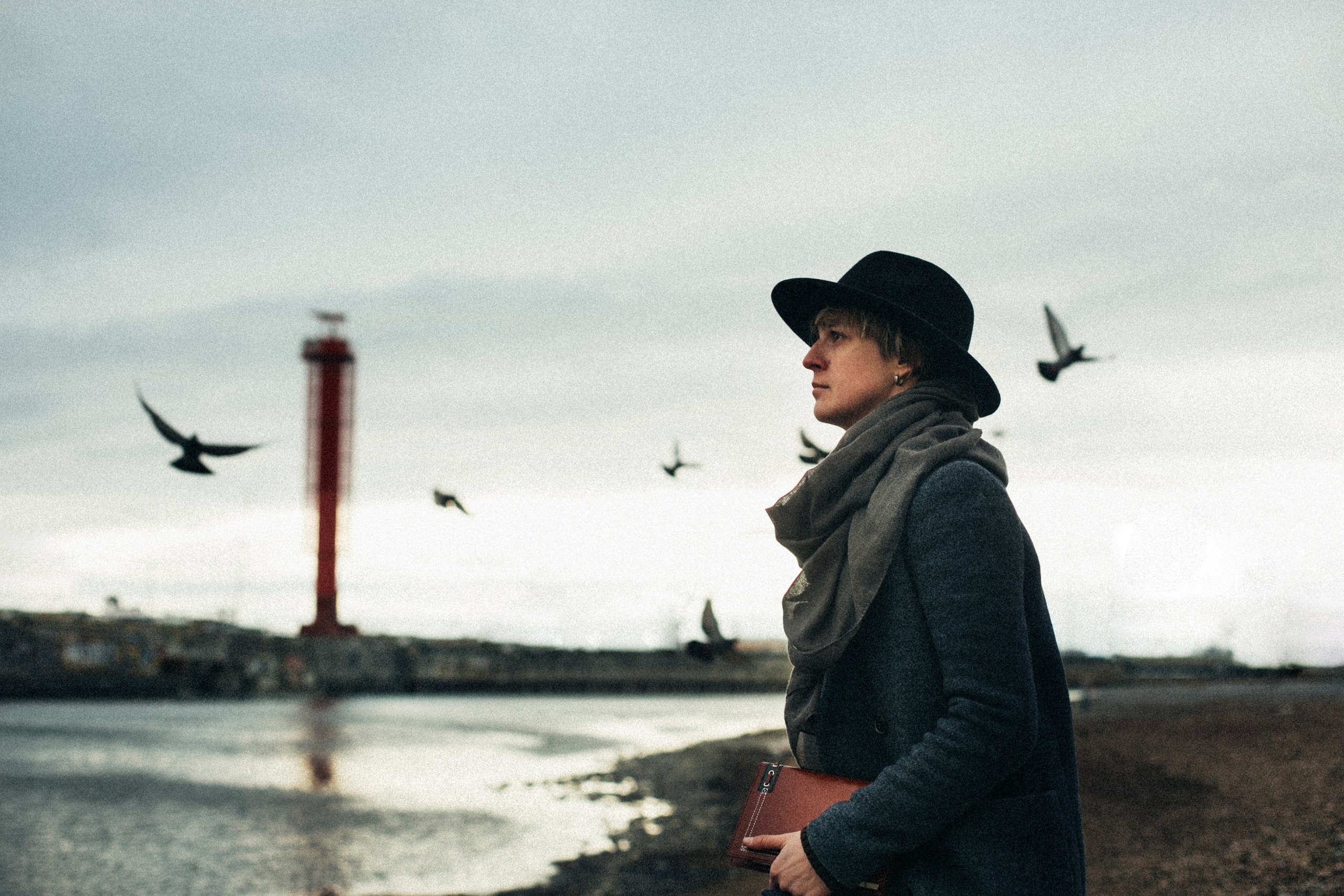 Атмосферный кадр на побережье, снятый фотографом из Сочи Галиной Скорик. Человек в пальто, шарфе и шляпе смотрит вдаль на фоне моря, летящих чаек и красного маяка.