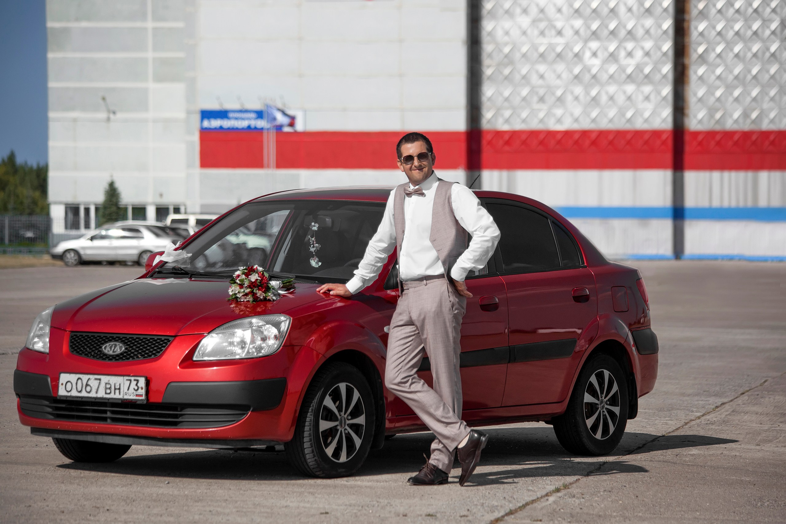 Портрет жениха у красной машины на фоне стены аэропорта в городе Ульяновск