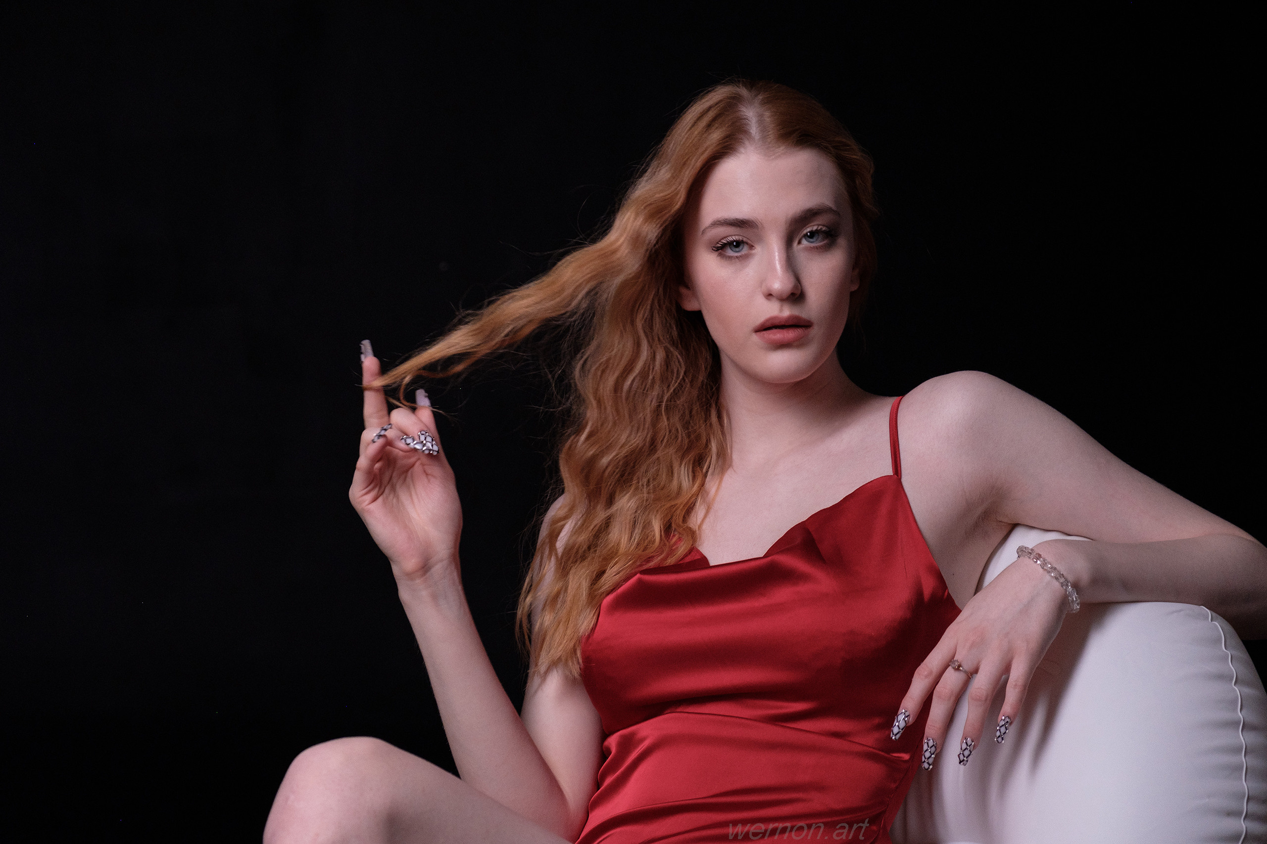 Блондинка в красном платье сидит в белом кресле и смотрит в камеру.