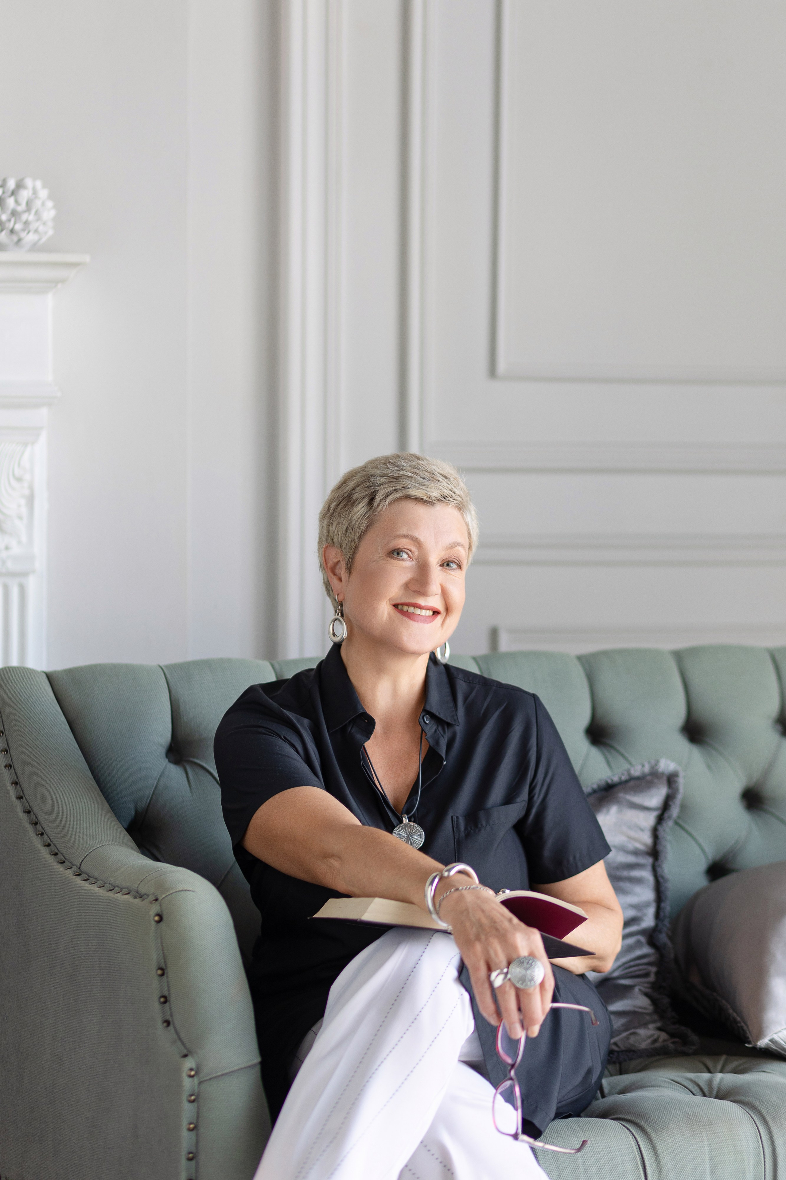 Женщина сидит на диване, облокотившись на подушку и улыбается.