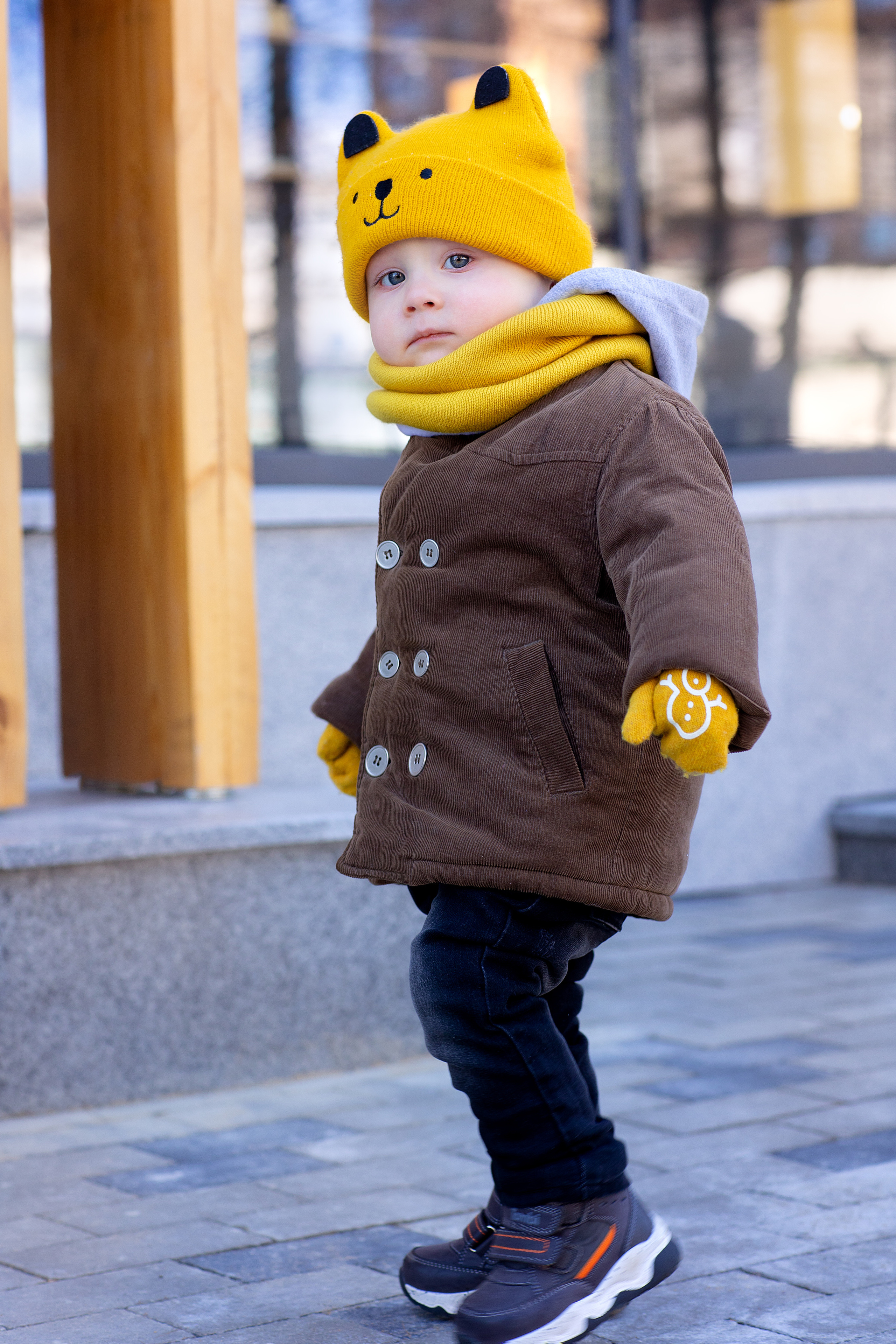 Мальчик в пальто и желтой шапке гуляет по городу на фоне витрин магазинов