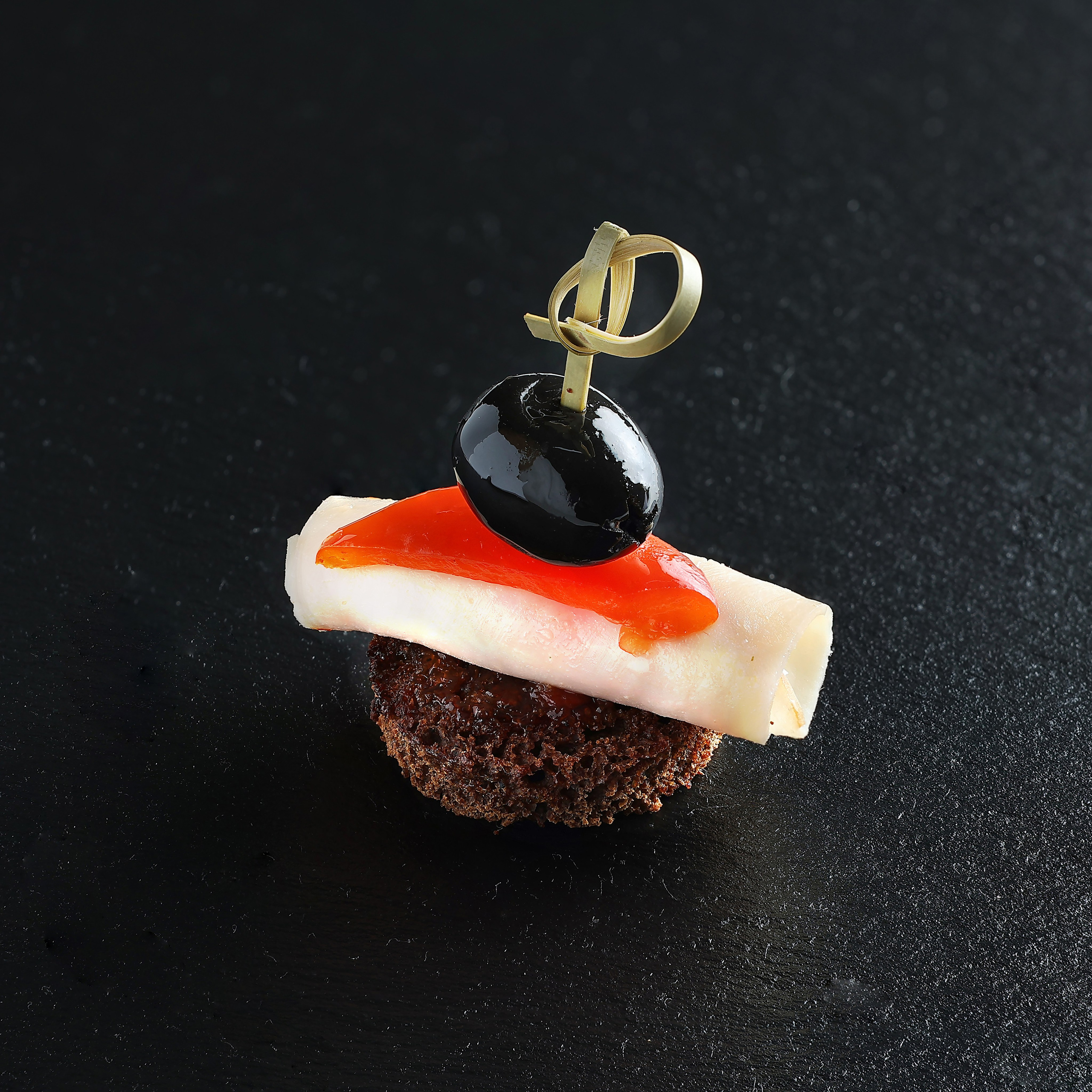 Графичная геометрия вкуса на выразительном тёмном фоне. Мини-закуска на бородинском хлебце с ветчиной, красным болгарским перцем и чёрной маслиной, скреплённая шпажкой, на тёмном фоне.