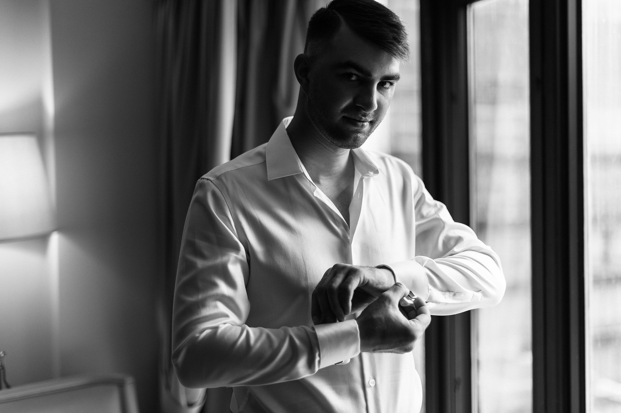 Жених застегивает рубашку возле окна в отеле.