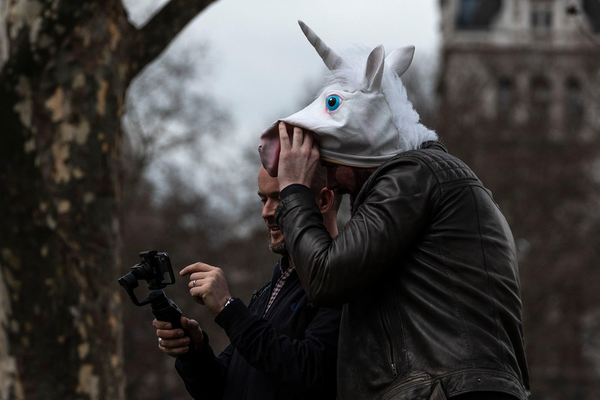 Два человека в Лондоне: один держит камеру на стабилизаторе, другой надевает маску единорога, позируя для съемки в парке.