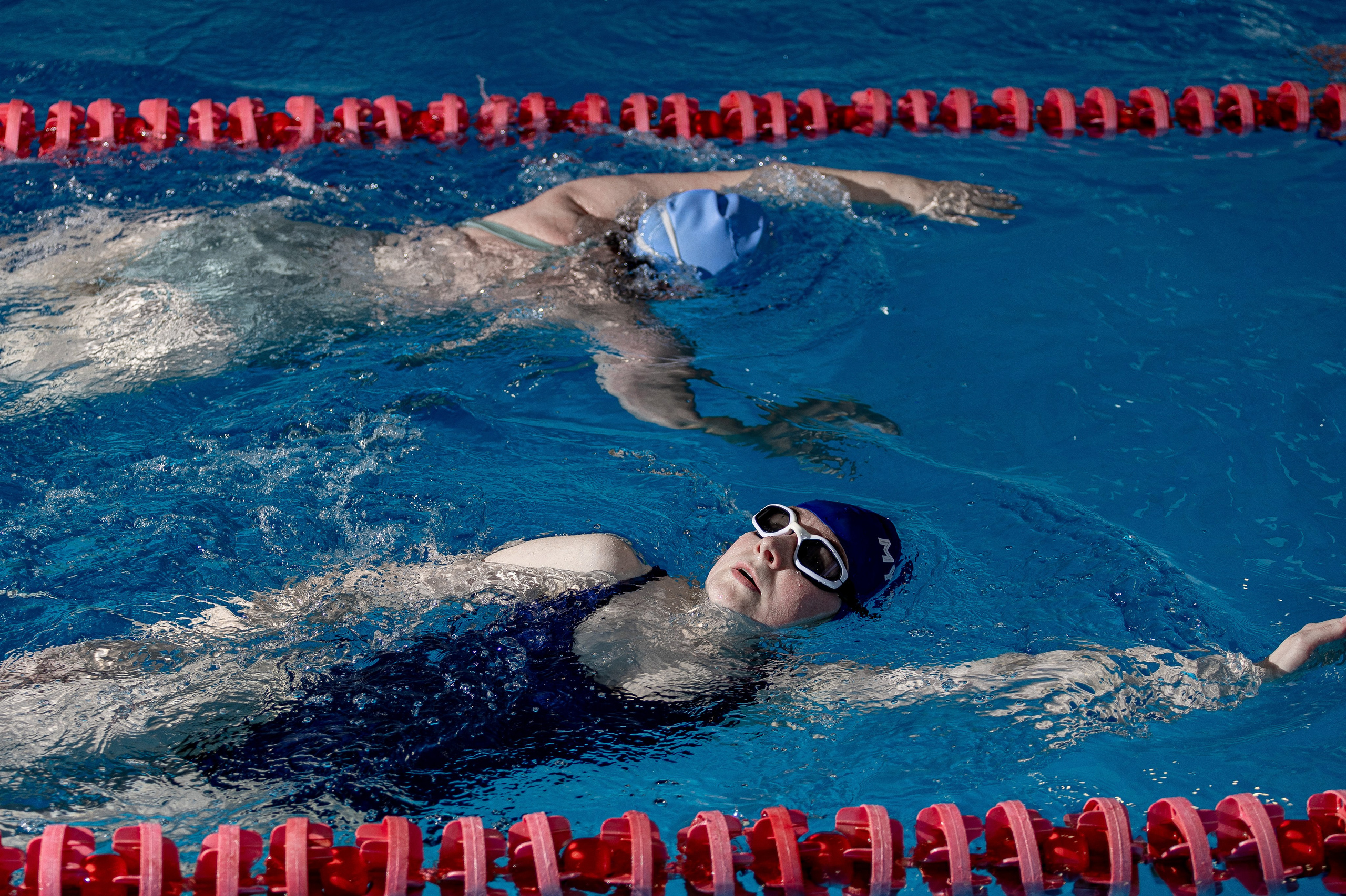 Спортсмены в купальных костюмах плывут кролем в бассейне, экипированы шапочками и защитными очками, на фоне — красные разделители дорожек.
