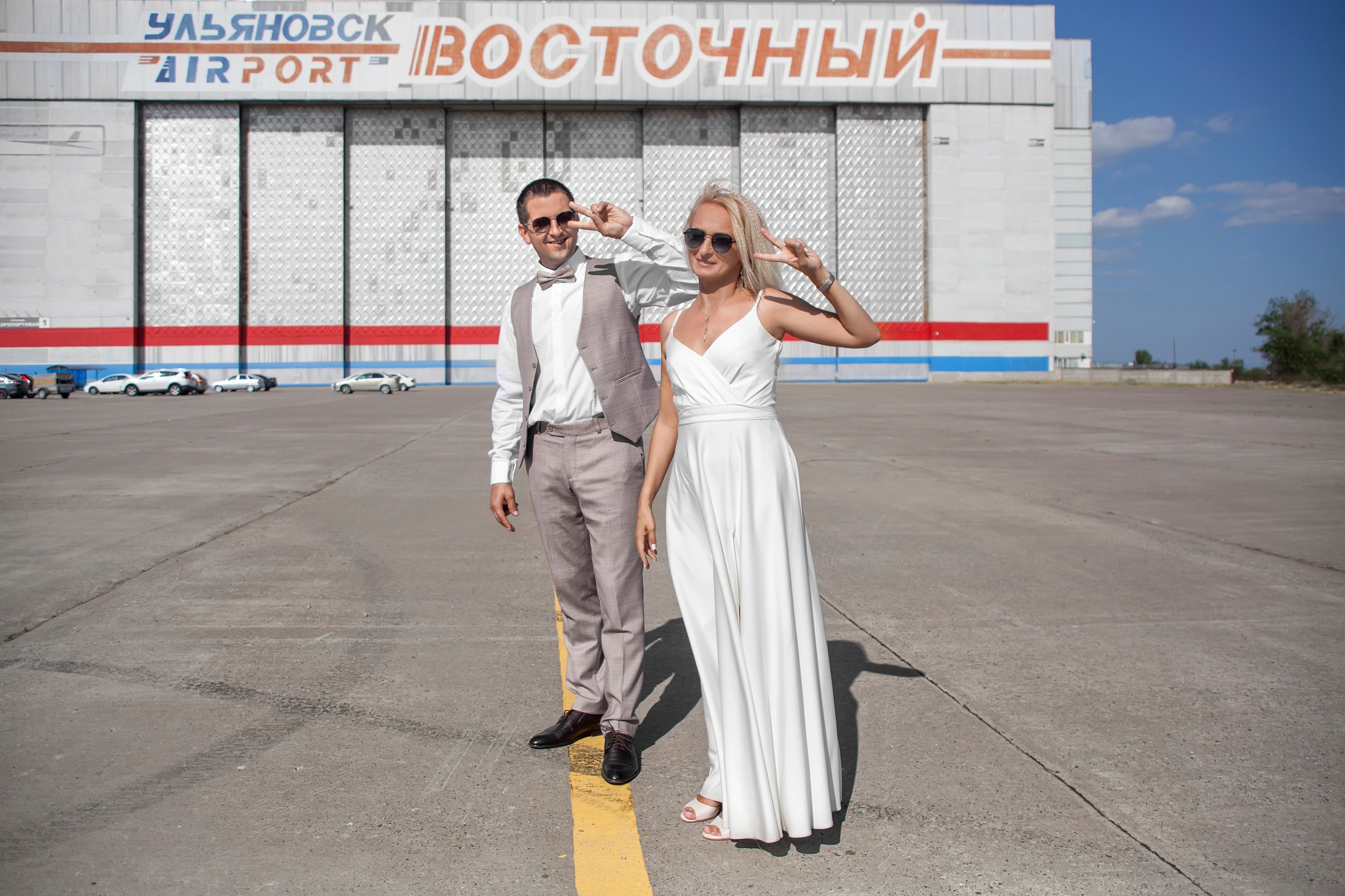 Жених и невеста делают одинаковый знак руками на фоне здания аэропорта - фотограф Янкаускене Наталья