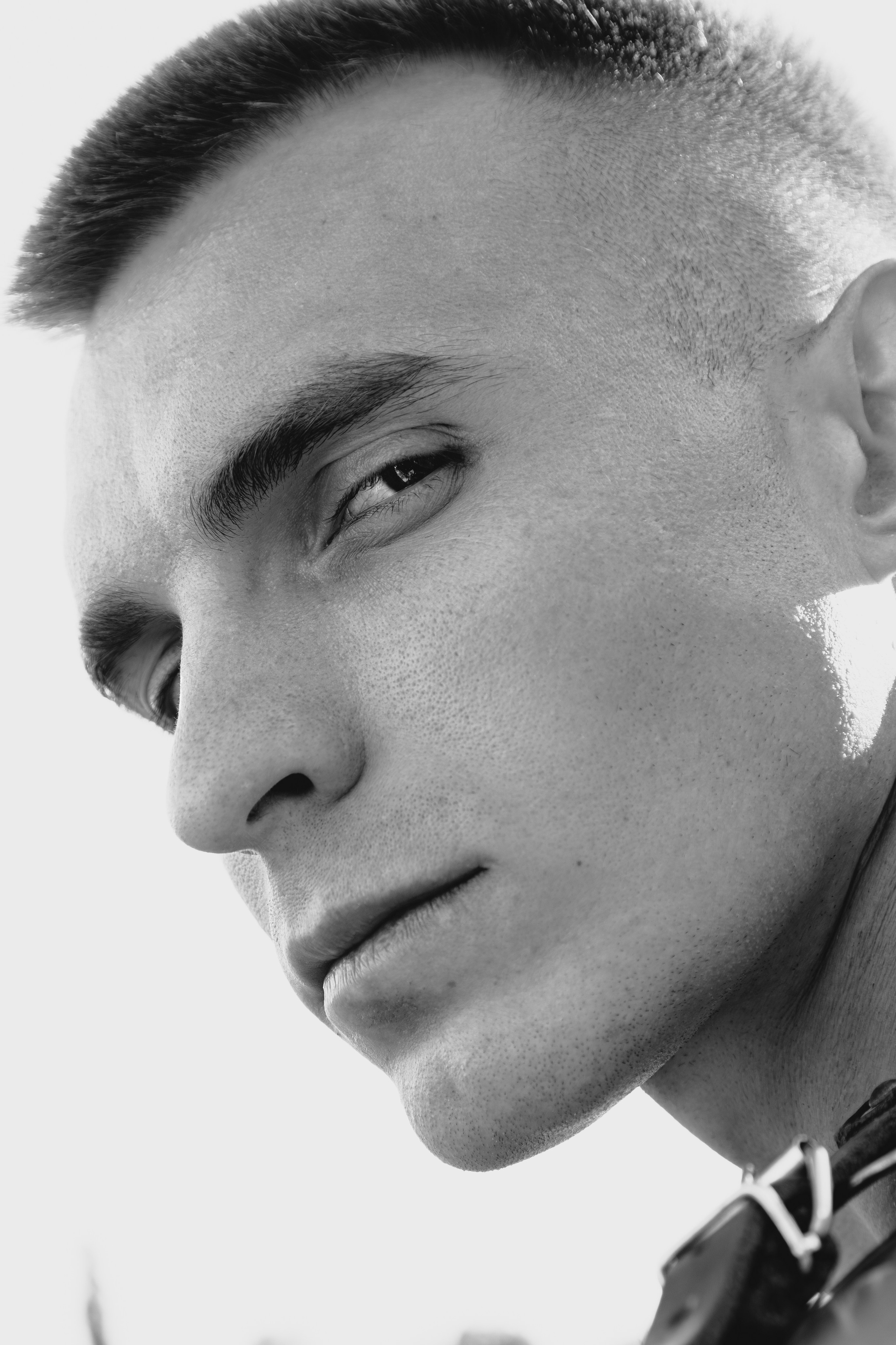 Сдержанная сила: мужской портрет в монохроме — крупный план, Ростов‑на‑Дону. Мужской портрет в ч/б: крупный план лица, выразительный взгляд, минимализм — фотосессия в Ростове‑на‑Дону. Крупный план мужского лица в чёрно‑белой гамме: скульптурные черты, лёгкая асимметрия взгляда, нейтральная полуулыбка; минималистичный светлый фон, акцент на фактуре кожи. Ростов‑на‑Дону. Молодой мужчина в профиль, взгляд чуть опущен и направлен в сторону; выражение лица спокойное, собранное, лёгкая полуулыбка; короткая стрижка с плавным переходом длины, чёткие контуры висков; кожа гладкая, с естественной фактурой и лёгкой щетиной; брови густые, дугообразные; нос прямой, губы сомкнуты, линия подбородка чёткая; на шее — фрагмент тёмного кожаного ремня с металлической пряжкой; фон — чистый, равномерно освещённый, без деталей; чёрно‑белая палитра, высокий контраст, мягкий направленный свет, фокус на глазах, скулах и линии челюсти.
