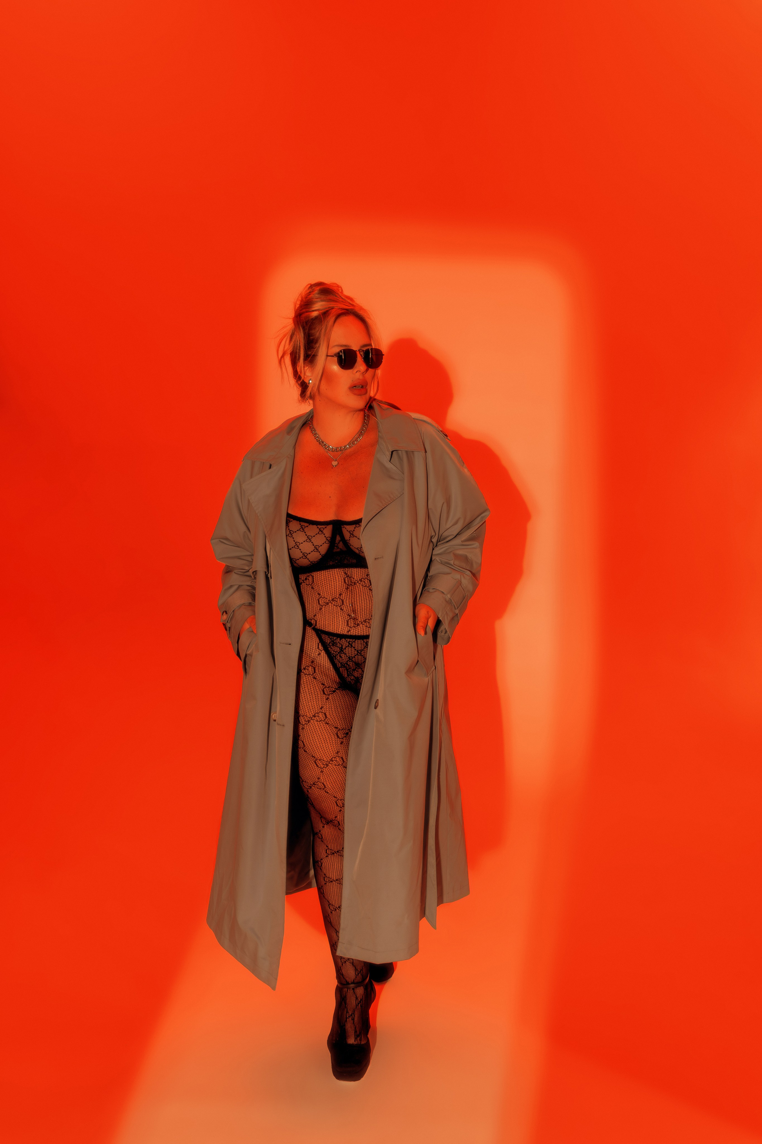 Эффектный fashion‑кадр с продуманным балансом дерзости и элегантности: насыщенный оранжево‑красный фон работает как активный художественный инструмент — он усиливает энергетику образа и делает снимок мгновенно узнаваемым. Центральный приём — контраст фактур и слоёв: лаконичный бежевый тренч в расслабленной подаче (руки в карманах, лёгкая динамика шага) противопоставлен выразительным сетчатым фактурам белья и чулок, что создаёт интригу и многослойность.Дополняют образ солнцезащитные очки в ретро‑силуэте — они добавляют загадочности и завершают стилистическую формулу «уверенная непринуждённость». Аккуратное колье‑цепочка служит деликатным акцентом, не перетягивая внимание с общей композиции.Такой подход идеально подходит для:лукбуков брендов одежды и белья, где важна подача фактур;контент‑съёмки для соцсетей в стиле fashion и lifestyle;портфолио моделей плюс‑сайз с акцентом на уверенную подачу;рекламных кампаний с яркой цветовой доминантой.