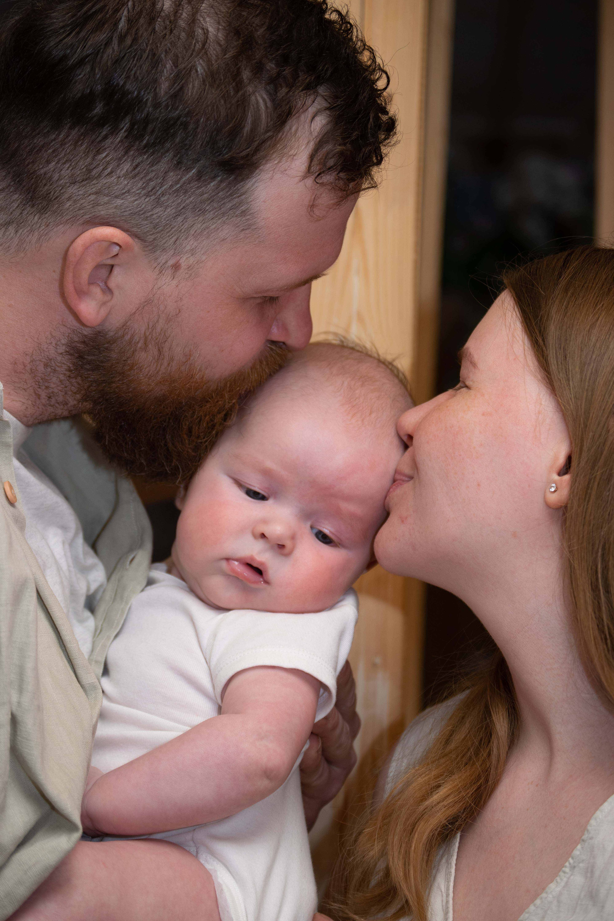 Семейная фотосъемка молодой семьи. Родители целуют малыша.