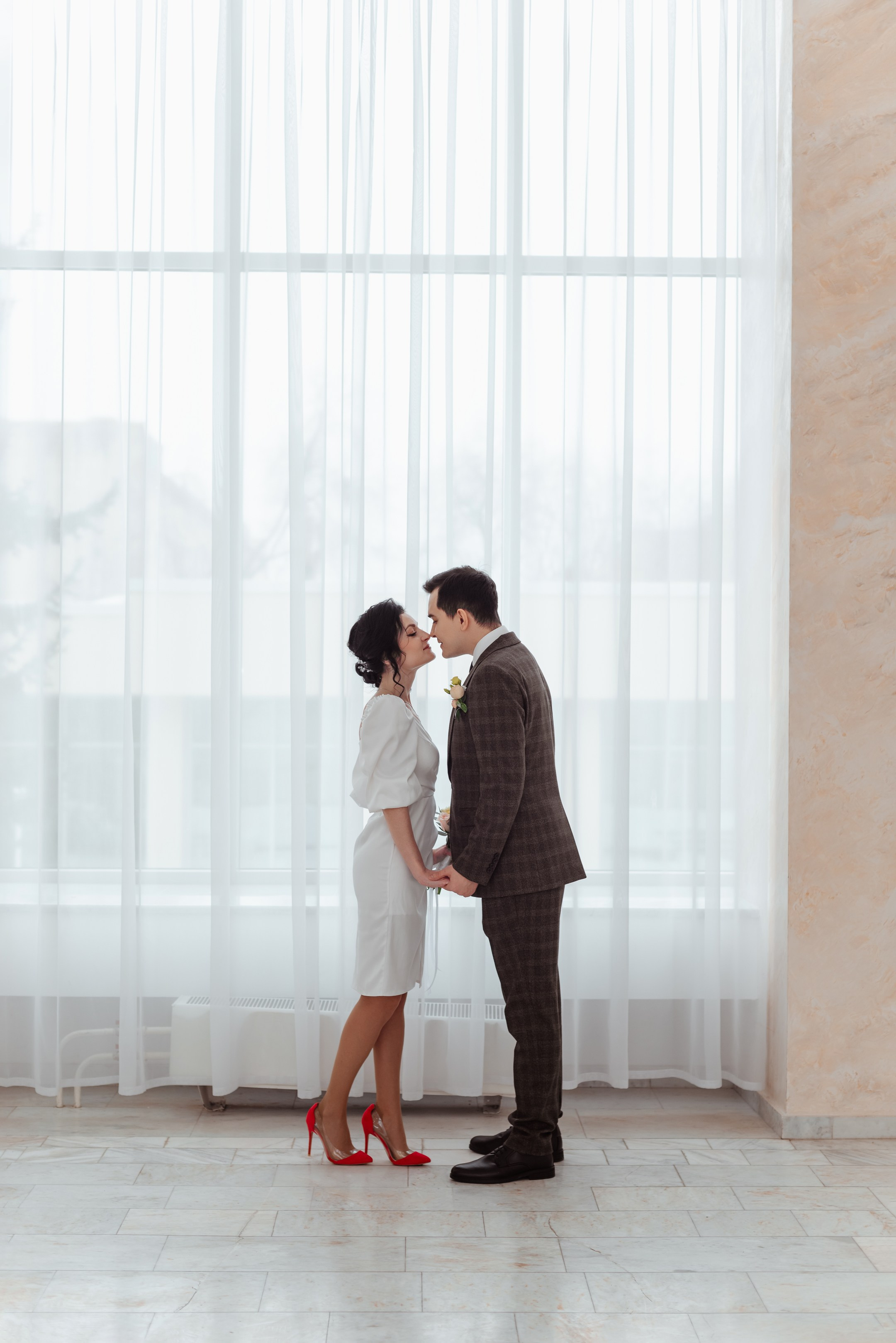 Молодые супруги целуются в холле ЗАГСа, невеста в белом мини-платье, жених в коричневом костюме-тройке.