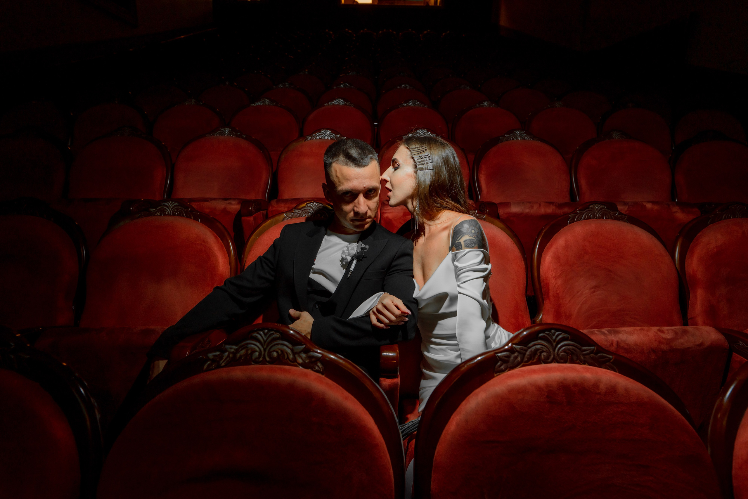 Жених смотрит в кадр, а невеста целует его в щеку и сидят в темном зале театра Андрея Миронова
