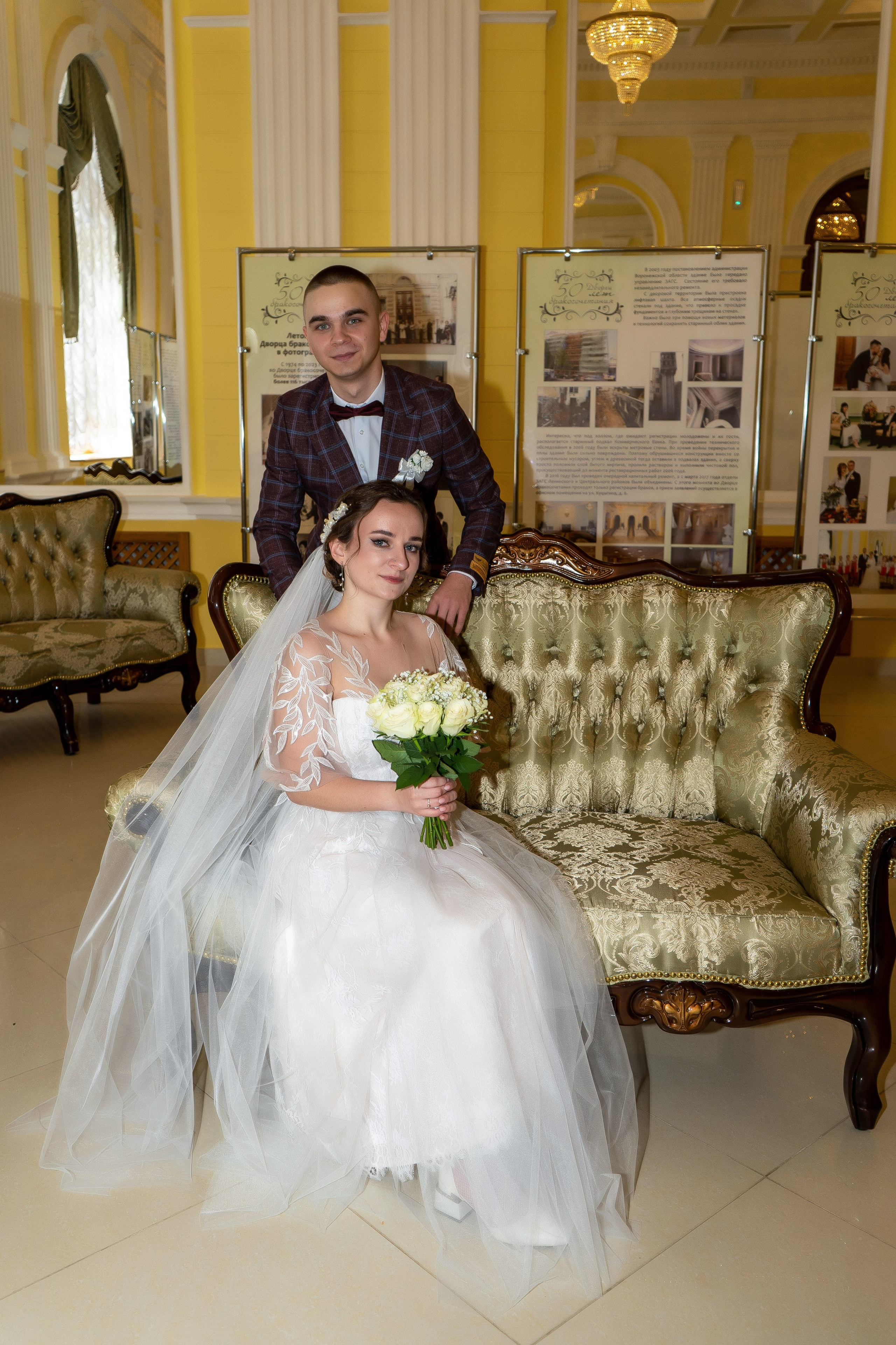 Невеста в свадебном платье с длинной фатой сидит на диване с букетом роз, жених обнимает сзади в Воронеже