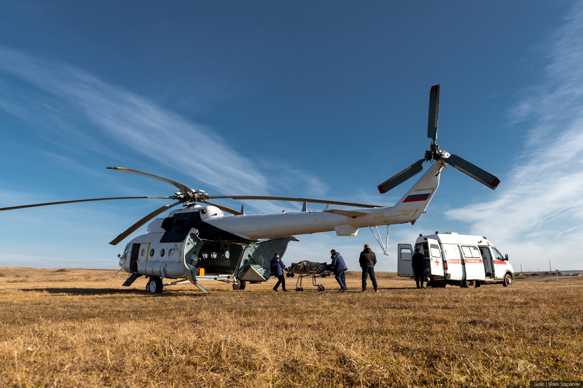 Вертолёт санитарной авиации Ми-8 с открытыми створками хвостового отсека в поле. Медицинские работники перемещают пациента на каталке к реанимобилю. Фотограф — Slava Stepanov.